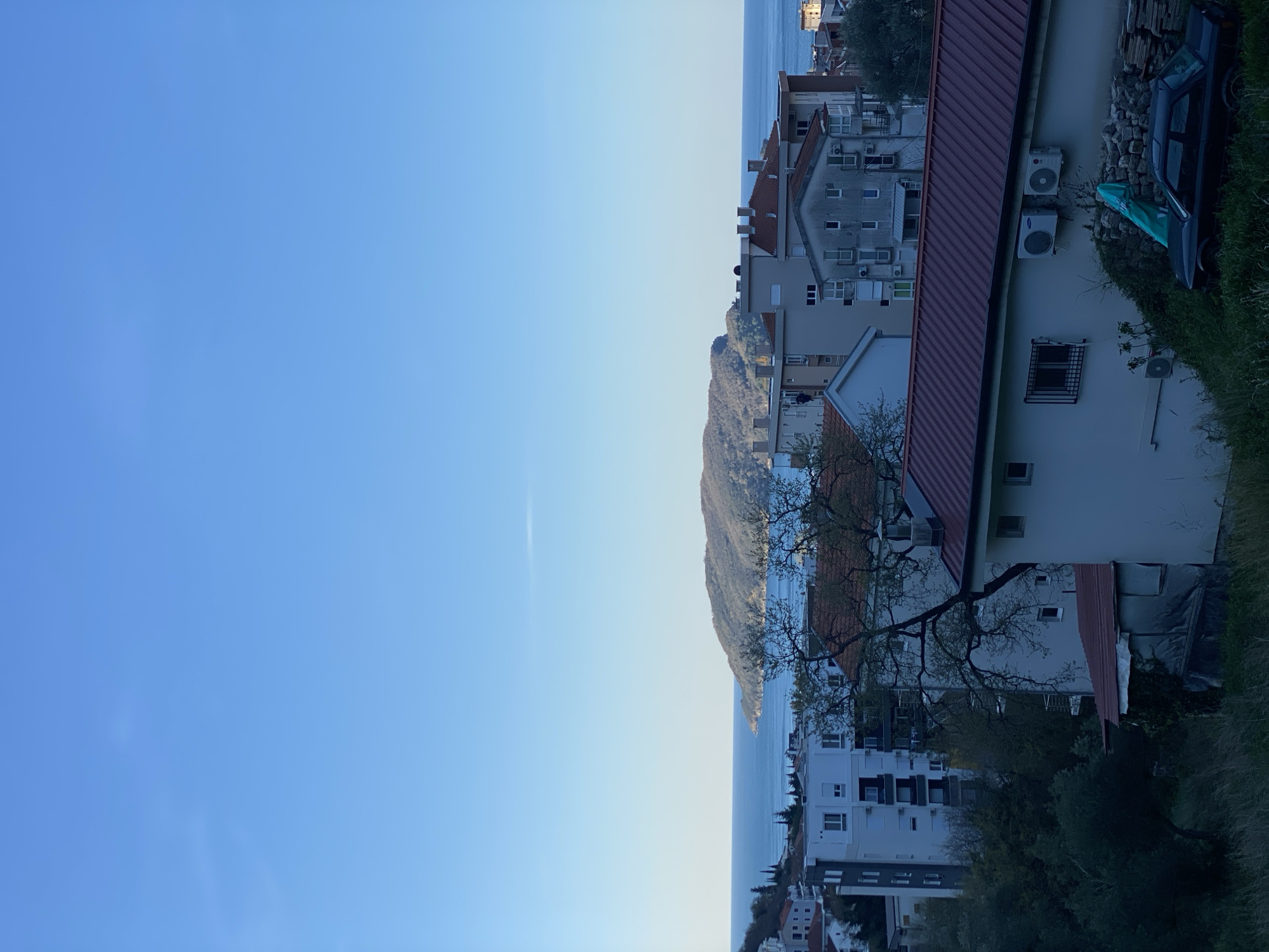 The image depicts a daytime scene in Budva, Montenegro. The sky is clear blue. From an elevated vantage point, a partial view of the Adriatic Sea and a mountainous peninsula jutting into the water is seen. Closer to the viewer are dense residential buildings, characterized by white facades, red roofs, and multiple stories. Air conditioning units are affixed to some buildings. A deciduous tree, partially in bloom, stands near the buildings. Adjacent to one of the buildings is a pile of rocks, a dark-colored car, and some vegetation. The orientation suggests the photograph was taken vertically.