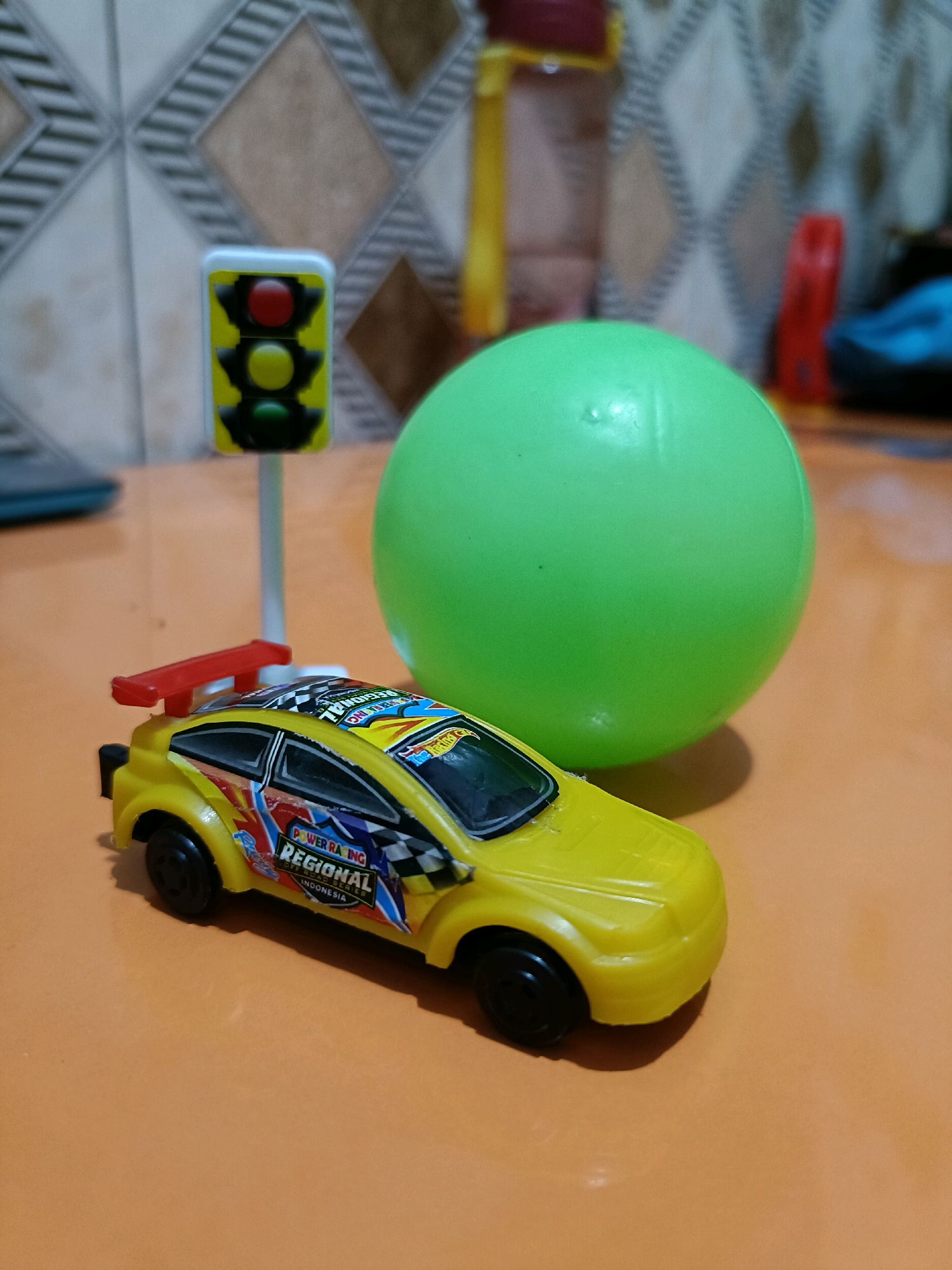 In Candi Wates, Indonesia, a toy traffic light with red, yellow, and green lights stands on a white pole. Beside it, a bright green ball rests on an orange surface. In the foreground, a yellow toy race car with colorful decals, including "POWER RACING REGIONAL INDONESIA" and a checkered flag, is positioned on the same orange surface. The background features a patterned wall and other indistinct objects.