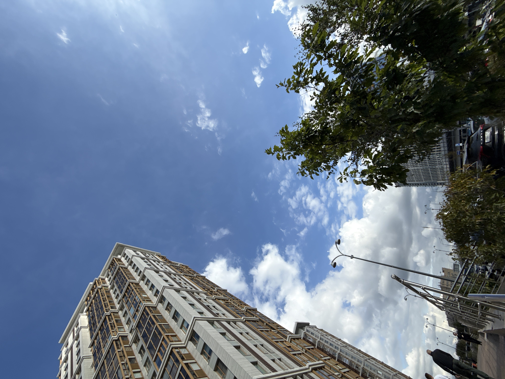 The image depicts a street scene in Moscow, Russia, featuring a tall, modern apartment building with a distinctive geometric facade and numerous windows. The building is angled upwards, dominating the left side of the frame. The sky is a bright blue with scattered white clouds, and the sun is visible, casting light and creating a slight glare. In the foreground and mid-ground, there are trees with green foliage, some of which are partially obscuring the view of the street and other buildings. A street light with two lamps is prominent in the mid-ground. Several people are visible walking on a sidewalk, some with their backs to the camera, dressed in casual attire. A metal railing runs along the side of the sidewalk. In the background, other buildings, including a glass-fronted structure, and cars on a road can be seen. The overall impression is a contemporary urban environment on a clear day.