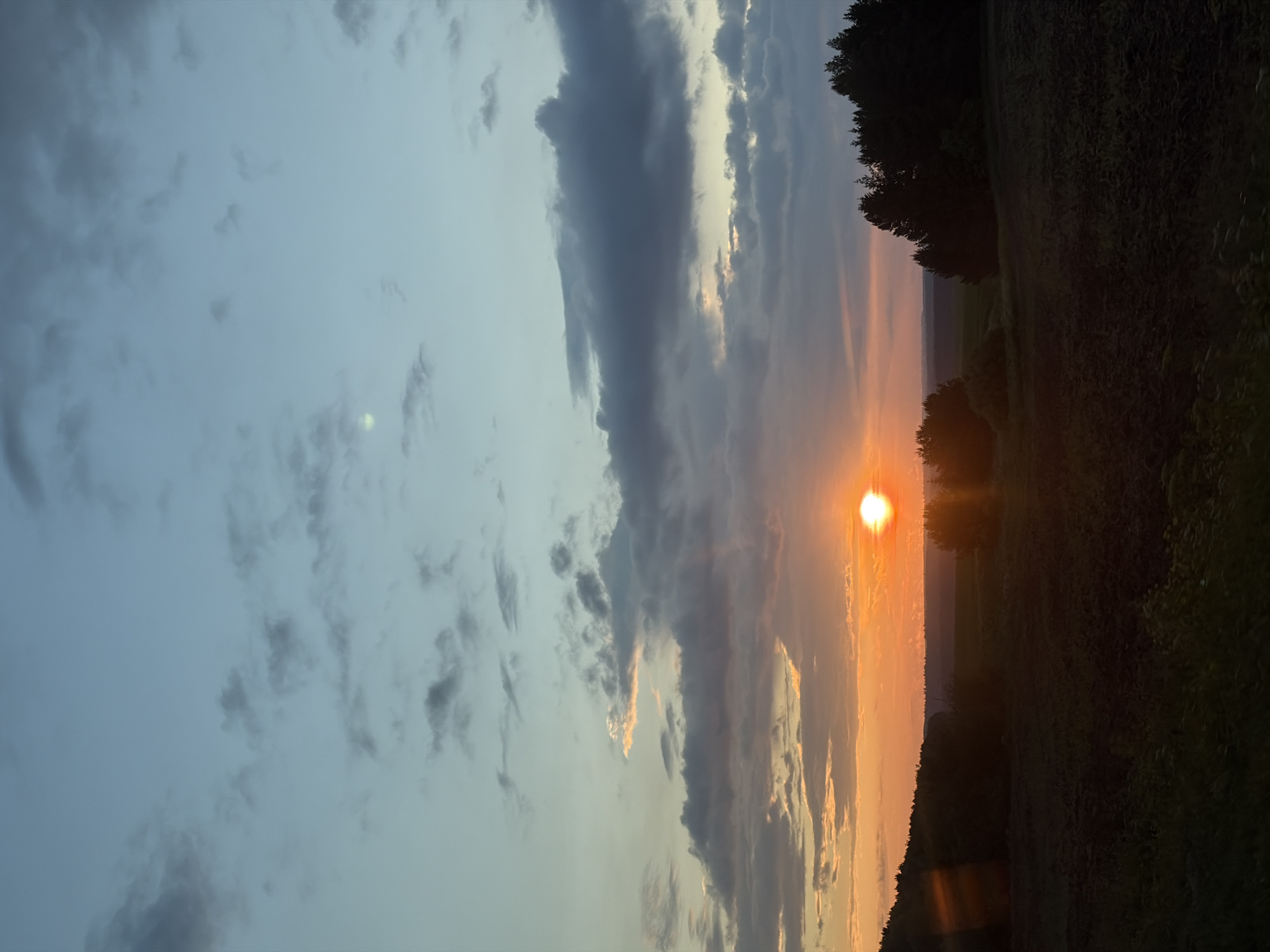 A vertical, full shot captures a dusk scene in Lukintsy, Russia. The sky transitions from a light blue at the top to a warm orange glow near the horizon, where the sun is setting. The sun is a bright, distinct orb positioned slightly to the right of the frame's center. Clouds are scattered across the sky, some thin and wispy, others darker and more substantial, with the lower clouds catching the orange and pink hues of the sunset.
Below the sky, a dark, silhouetted landscape unfolds. A line of dense evergreen trees forms a dark mass along the right side of the frame, extending inwards. To the left, rolling hills are visible, also covered in dark foliage. In the foreground, a field stretches across the bottom of the image. The field appears to be cultivated land, with dark soil and some low-lying vegetation, largely in shadow. The overall impression is one of natural beauty and tranquility as day transitions into night. There are no people or animals visible in the image.