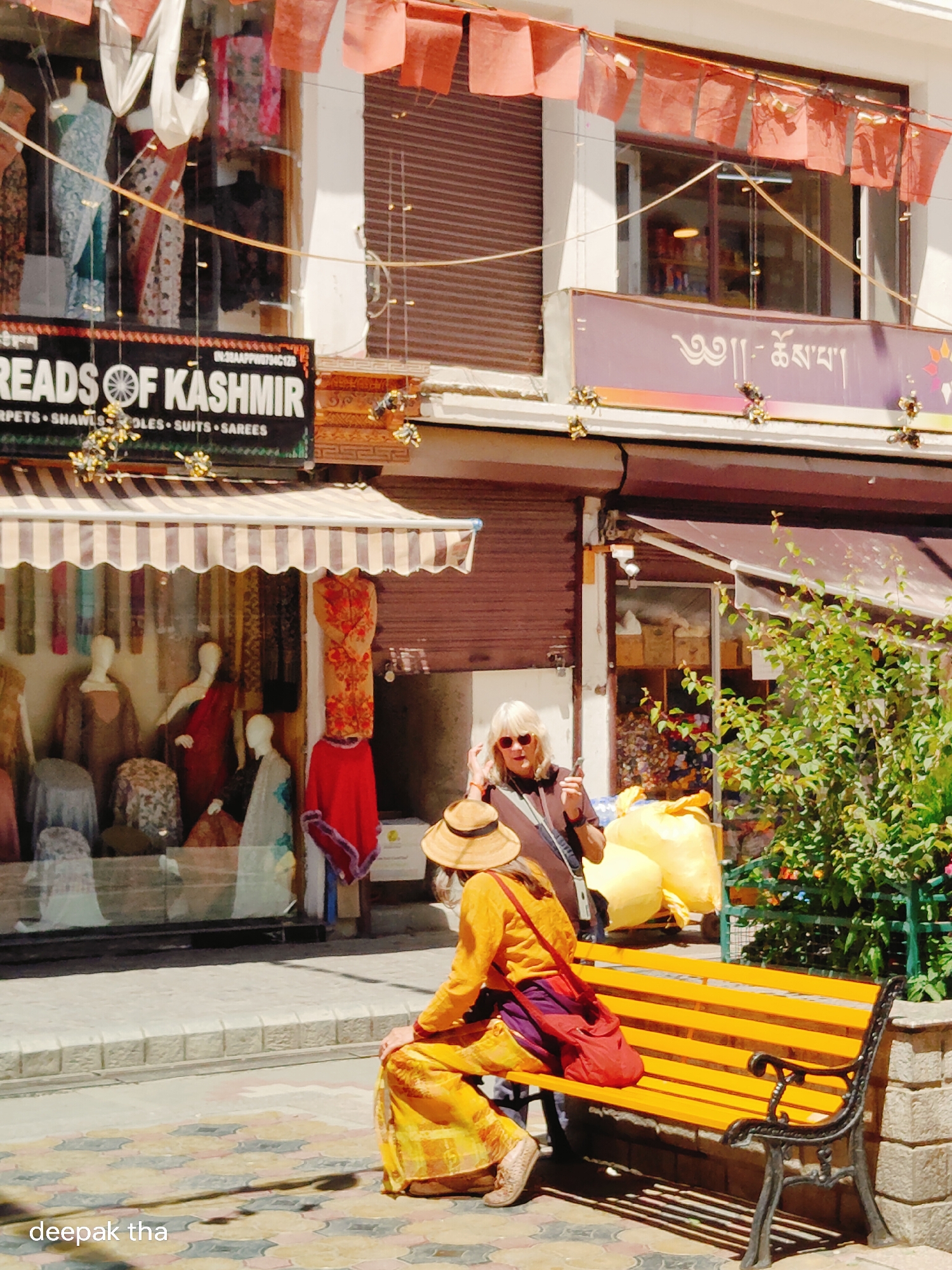 The image captures a street scene in Leh, India, likely in the market area. In the foreground, a person is seated on a bright yellow bench, facing away from the camera. They are wearing a straw hat and a traditional yellow and orange outfit, with a red bag resting on their lap. To the left of the bench, a woman with blonde hair and sunglasses is standing, holding a mobile phone and looking towards the right. Behind them, a storefront displays clothing on mannequins and is adorned with red prayer flags overhead. The shop's signage includes the text "THREADS OF KASHMIR" and what appears to be Tibetan script above. The street is paved with patterned tiles, and a green bush grows to the right of the bench. The overall impression is a sunny day in a vibrant, bustling street.