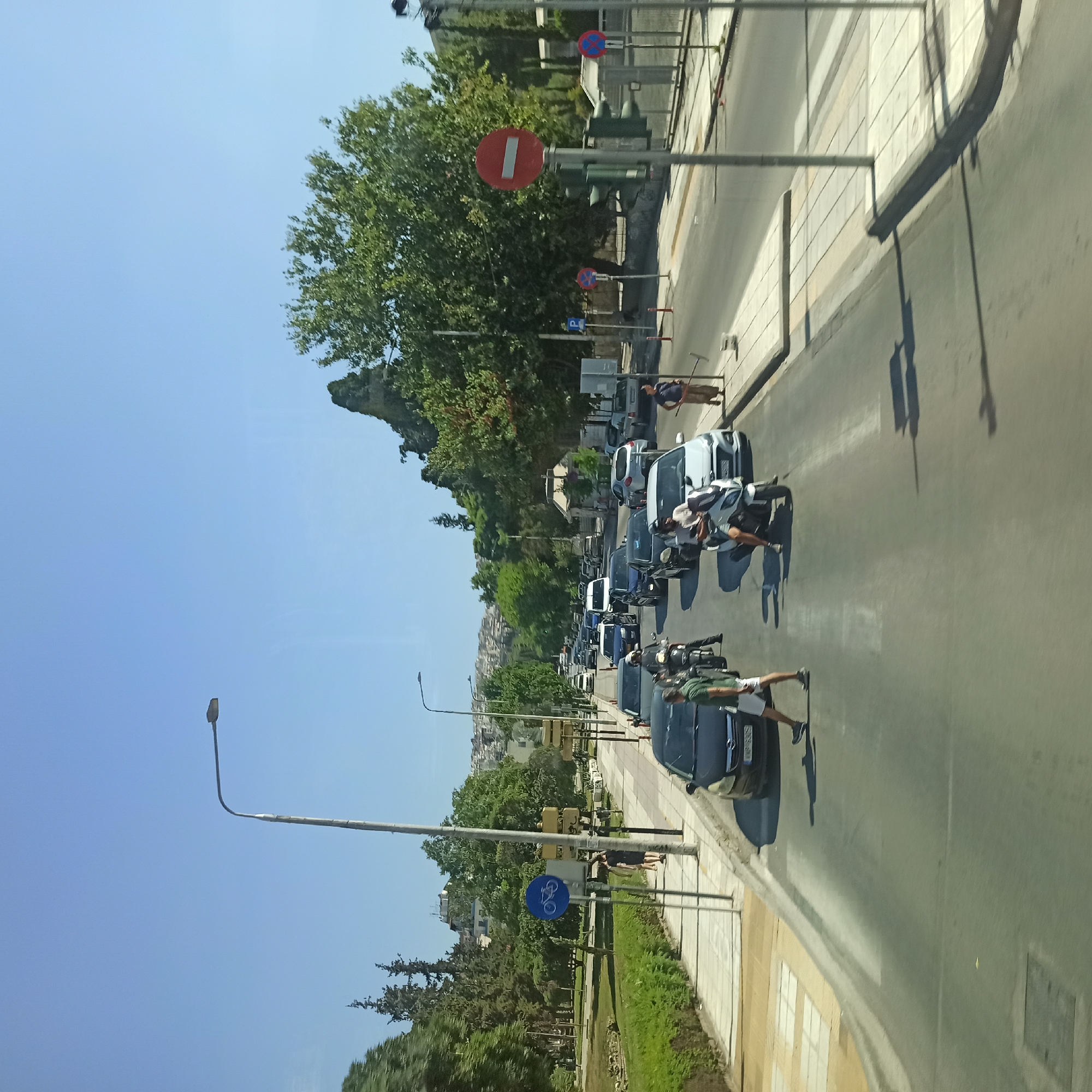 The image depicts a street scene with moderate traffic congestion on a sunny day. The viewpoint is from within a vehicle, looking forward. On the left side of the road, a pedestrian walkway runs alongside a green area with trees. A bicycle lane is indicated by a blue sign with a white bicycle icon. Further down the walkway, two people are visible, one walking and the other standing.

The road itself has multiple lanes of traffic, with cars and motorcycles present. Several vehicles are stopped or moving slowly, suggesting a traffic jam. A person is crossing the street at a pedestrian crossing, casting a shadow on the asphalt. On the right side of the road, there are more pedestrian walkways and traffic signs, including a red circular sign with a white horizontal bar, indicating "Do Not Enter". A traffic light is also visible.

In the background, buildings are visible on a hillside, with dense foliage from trees obscuring some of them. The sky is clear and blue. The presence of a bicycle lane sign and the urban setting with buildings on a hill suggest a location within a city. Given the presence of specific street signs and architectural styles, this scene is likely from the city of Thessaloniki, Municipal Unit, Greece. The overall impression is of a typical urban environment experiencing daytime traffic.