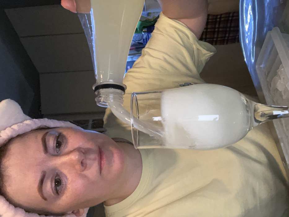 A woman with a pink headband pours a light-colored liquid from a clear bottle into a glass. The glass is nearly full of a cloudy white substance, possibly a blended drink. An ice cube tray is visible in the background. The woman wears a light yellow t-shirt. She looks directly at the camera, close-up, with a neutral expression.