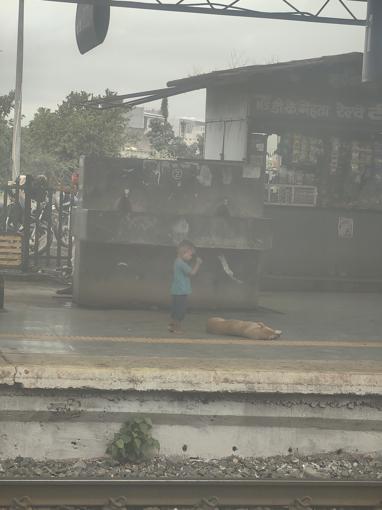 The image depicts a railway platform in Chhanera, India. A young child wearing a blue t-shirt and dark shorts stands near a concrete structure, holding what appears to be a mobile phone or small camera. A light brown dog is lying down on the platform in front of the child. In the background, there is a stall with various items displayed, possibly snacks or drinks. Behind the stall, trees and buildings are visible. The platform shows signs of wear, with some cracked concrete and debris. The sky is overcast.