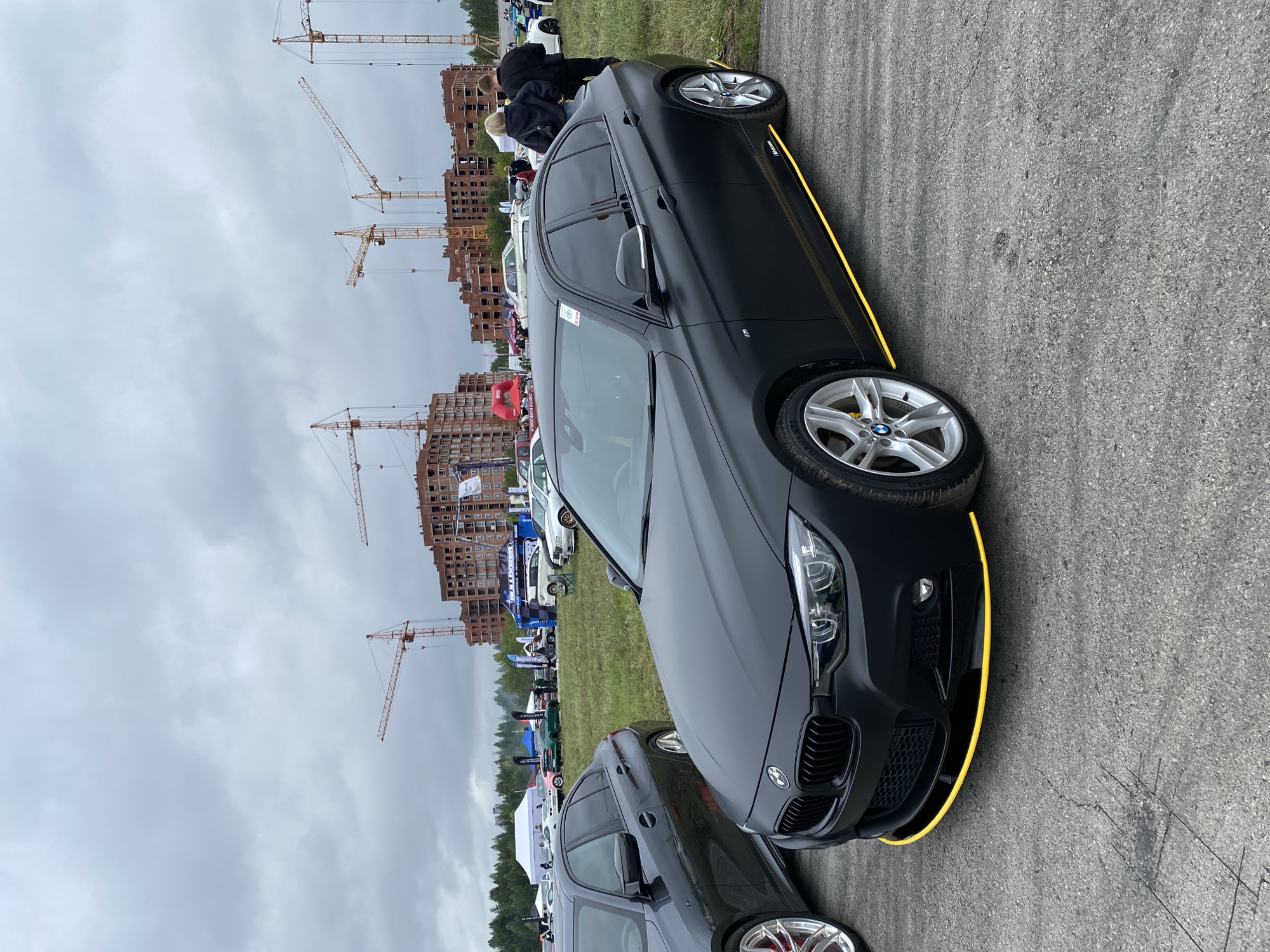 The image shows a matte black BMW sedan with a yellow stripe on its lower body kit, parked on an asphalt surface. The car has silver rims with yellow brake calipers. In the background, there are several unfinished brick buildings under construction, with numerous construction cranes visible in the sky. Various tents and other vehicles are also present, suggesting an outdoor event or exhibition. The sky is overcast with clouds. The scene appears to be an outdoor event or exhibition, possibly a car show, in a developing urban area, with the construction site indicating a location in the city of Perm, Russia.