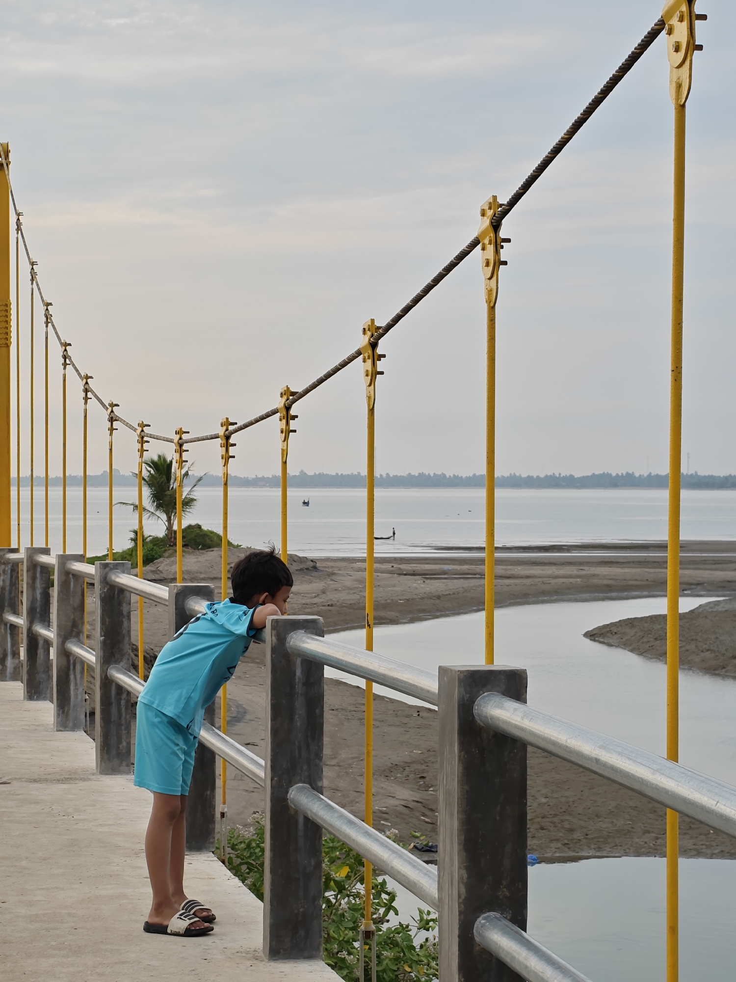 A young child, dressed in a blue t-shirt and shorts, leans on a railing overlooking a wide body of water. The child is standing on a concrete walkway, likely part of a bridge, with bright yellow vertical supports and thick cables spanning overhead. The railing consists of concrete posts and horizontal metal bars. The child's gaze is directed towards the water and the sandy shore beyond.
The scene appears to be set during the day, with an overcast sky suggesting possibly cloudy or hazy weather. The water is calm, and a small boat is visible in the distance, along with a treeline along the far shore. The foreground shows a sandy area with a small river or inlet winding through it. A lone palm tree stands on a small mound of land to the left, adding a tropical element to the landscape.
The overall mood of the image is one of quiet contemplation, with the child appearing absorbed in the view. The location appears to be a coastal or riverside area in Lhokseumawe, Indonesia, suggested by the presence of palm trees, the type of vegetation, and the architecture of the bridge. No visible text is present in the image.