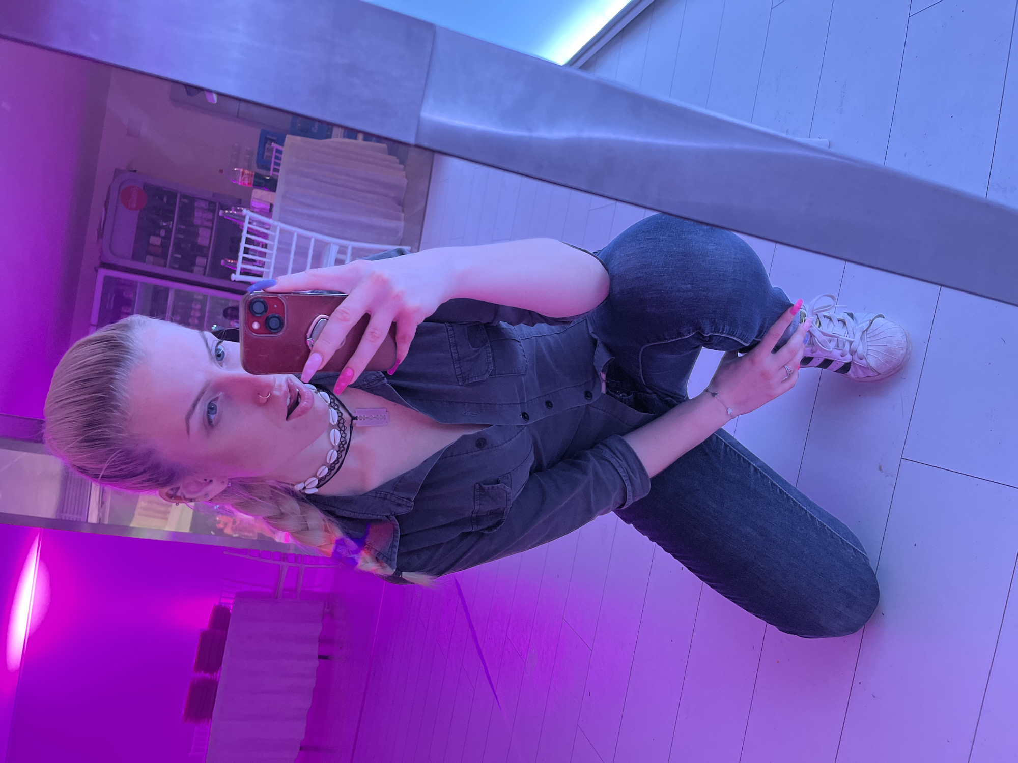 A young woman is taking a selfie in front of a mirror. She is kneeling, wearing jeans, a dark button-down shirt, and white Adidas sneakers. She has pink nail polish and is holding a phone with a maroon case. The reflection in the mirror shows a display of items behind her, possibly a refrigerator and some shelving units. The overall lighting is a mix of blue and purple hues. The location is likely indoors, in a commercial setting in Rakovski, Bulgaria.