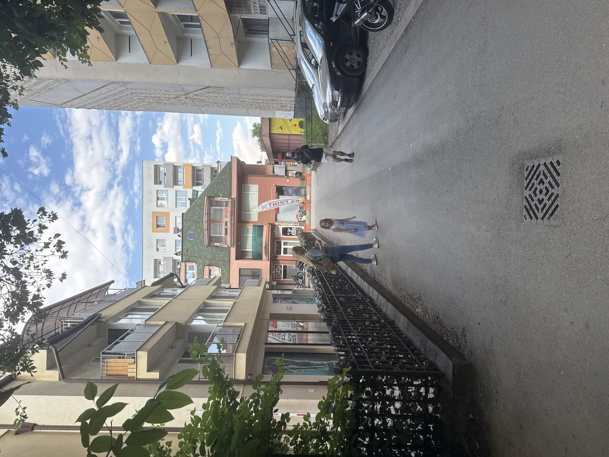 This image shows a street scene in the city of Sofia, Bulgaria. A woman in jeans and a green shirt, carrying a backpack, walks with a young girl in a light blue dress. A man wearing shorts and a black t-shirt with a black bag slung across his chest walks away from them down the street. The street is lined with buildings of various architectural styles and colors. On the left, a cream-colored building with balconies and a dark metal fence is visible. On the right, a taller, light beige building with protruding balconies and a textured facade stands next to parked cars. In the background, there are more apartment buildings, one with a green roof. The sky is blue with scattered white clouds. A metal grate is visible on the asphalt street in the foreground.