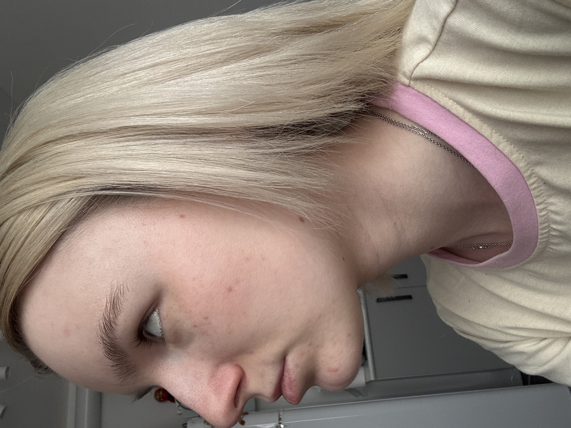 A side profile shot of a person with blonde hair, light skin, and light eyebrows is presented. The person is wearing a light-colored shirt with a pink trim around the neckline. A silver necklace is visible around their neck. The background is blurred, but suggests an indoor setting. There is no information in the image that specifies the location is in the city of Novoe Devyatkino, Russia.