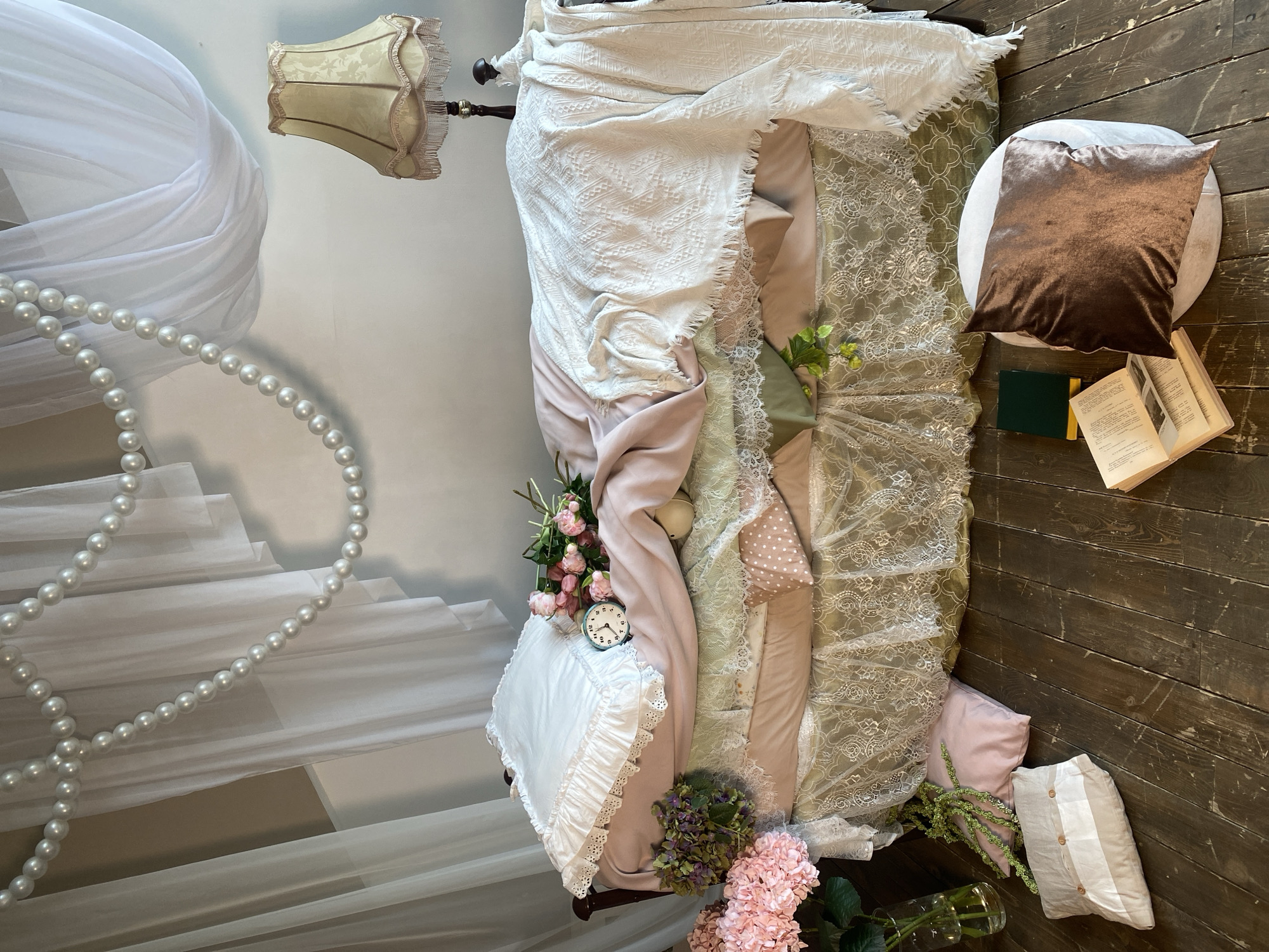 A bed with various linens and decorative items is the primary subject of the photo. The bed is draped with a white blanket and light pink bedding, along with lace elements. Several pillows are present, including a white one with lace trim and a brown velvet pillow on a white cushion near the head of the bed.
Floral arrangements are scattered around the bed. Other decorations include a vintage alarm clock, a lamp, and a string of pearls. On the floor near the bed, are open books and a green notebook.
The background features a wall with a staircase design partially covered by white fabric. The floor is made of dark wood planks. The photo's location is not identifiable from the image content. There is no information related to the city of Yekaterinburg, Russia.