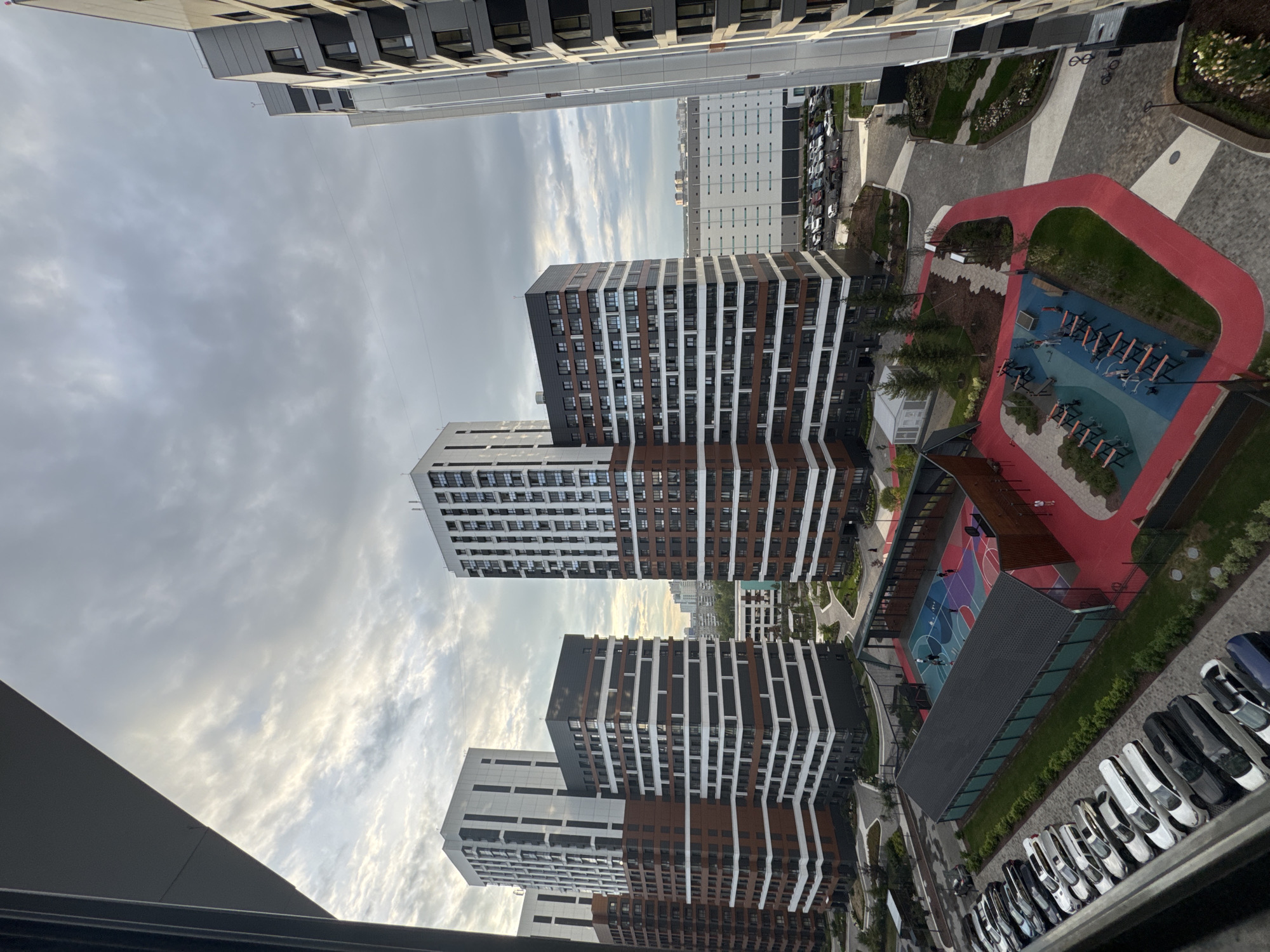The image depicts a modern residential complex in Novosibirsk, Russia. Several tall, multi-story buildings with a mix of brick and white facade elements dominate the skyline. The buildings feature numerous windows and balconies. In the foreground, a residential courtyard is visible, containing a children's playground with colorful equipment, a basketball court with visible players, an outdoor gym area, and pathways. Cars are parked in a designated lot to the left of the courtyard. The sky is overcast, with scattered clouds, suggesting a moderately cloudy day. The architecture suggests a contemporary urban development.