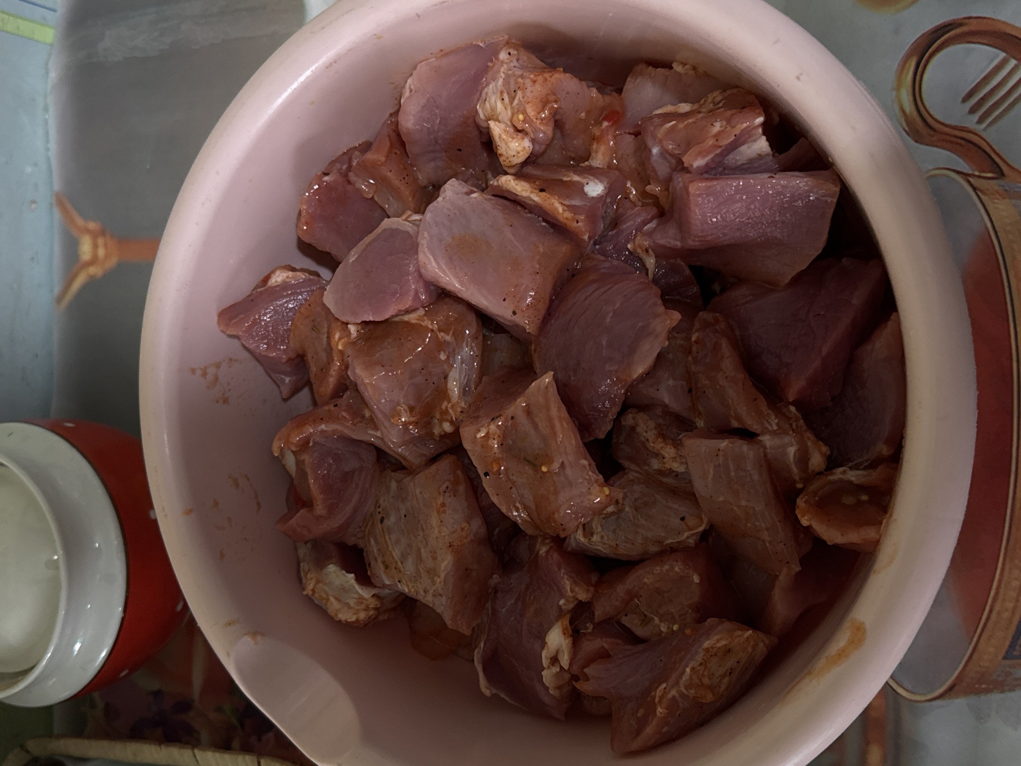 An overhead shot captures a light pink plastic bowl filled with cubed, marinated meat. The meat pieces are irregularly cut, exhibiting various shades of pink and reddish-brown, indicating they are raw and seasoned. A visible coating of dark specks, likely black pepper, and other reddish spices, along with a glossy sheen from oil or marinade, covers the meat.
The bowl itself is simple, with a slightly textured exterior. To the left of the main bowl, partially visible, is a red ceramic jar or pot with white polka dots and a white rim. The surrounding surface appears to be a patterned tablecloth or placemat. On the right side of the image, this surface features an ornate, stylized design of a fork and possibly a spoon in a golden-brown hue against a lighter background. Another portion of the background, particularly in the top left, is a muted, light blue-grey color, possibly a wall or another surface.
The scene suggests food preparation, with the meat likely marinating before cooking. No people are visible, and there is no discernible text in the image. The setting appears to be an indoor kitchen or dining area in Бородулино, Russia. The time of day and weather cannot be determined from the image.