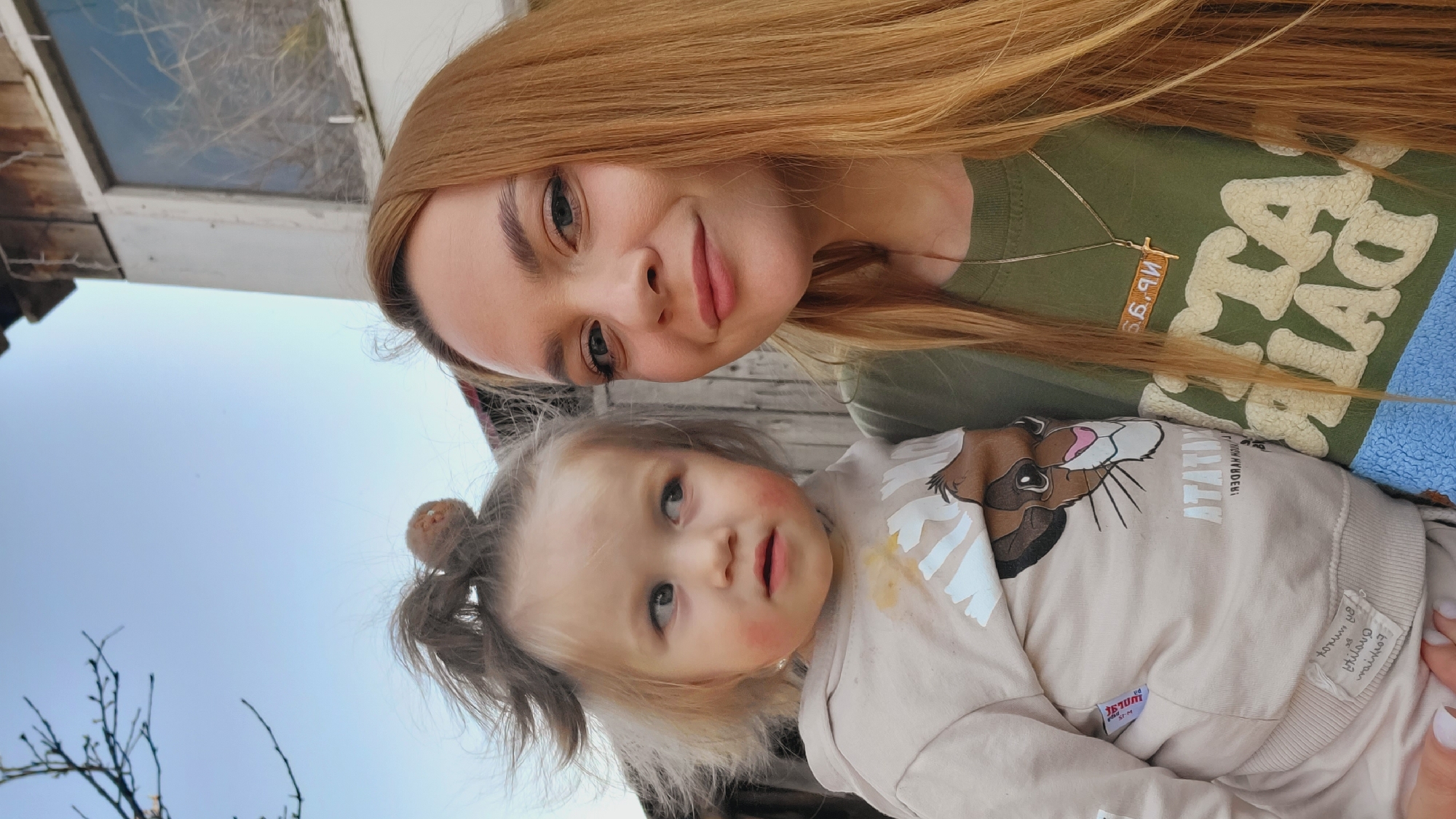 The image captures a selfie of a woman with her child. The woman has long, straight, light-brown hair and is wearing a green T-shirt with beige lettering. She also has a gold-colored necklace. The child is in the foreground and has blonde hair in a ponytail. The child is wearing a tan-colored sweatshirt with a cartoon lion on the front. There appears to be some food on the child’s shirt. The background features a wooden building with a window. The sky is visible above the building, along with bare tree branches. The location is potentially in Зипуново, Russia.