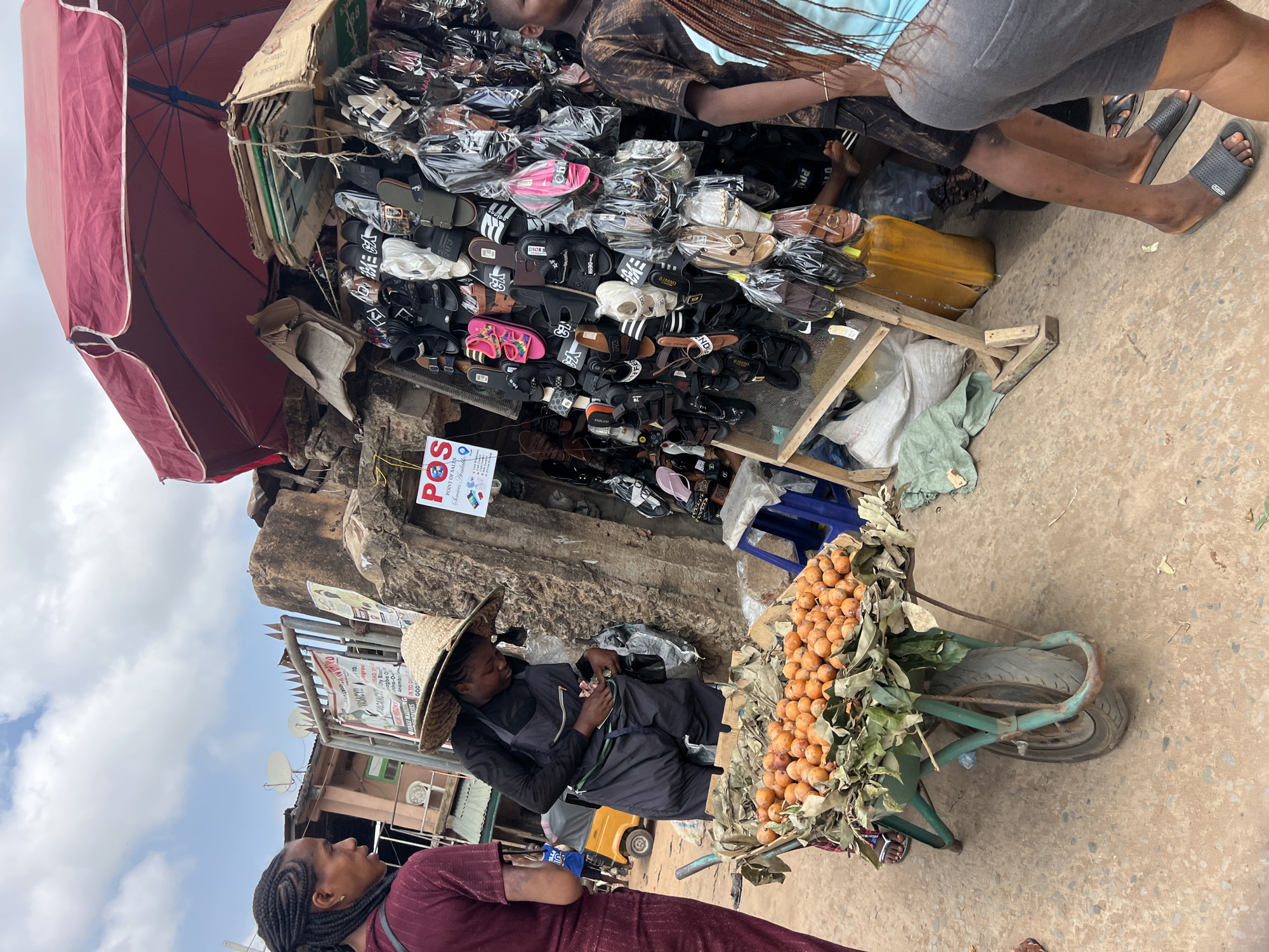 This image captures a vibrant street market scene in Nkpor, Nigeria, bustling with activity under a partly cloudy daytime sky. The primary focus is on two adjacent vendor stalls.
To the left, a large red market umbrella partially shades a shoe stall. The stall is laden with numerous pairs of sandals and slippers of various styles and colors, many individually packaged in clear plastic. Notable brands like "DIOR," "SIMMI," "ENDLESS," "ZARA," and "adidas" are visible on the footwear, suggesting a mix of popular designs, likely counterfeit in this informal setting. Several people are present around this stall, including a vendor or customer with braided hair wearing a light blue top and grey shorts, whose legs and arms are visible as they lean over the merchandise. Another person's legs, wearing black slides, are also seen near the stall.
To the right, a vendor sits beside a wheelbarrow filled with a pile of small, orange-colored fruits, possibly loquats or a local equivalent, nestled among green leaves. This vendor wears a straw hat and dark clothing, appearing to handle money. In the foreground, another person with braided hair and a maroon top is partially visible, looking towards the market activity.
The ground is a mixture of dirt and concrete. In the background, simple buildings with satellite dishes are visible, along with various signs attached to a wall. A red and white sign prominently displays "POS," "POINT OF SALES," and "Services Available," indicating financial transaction services. Other partially legible texts on signs include "VACANCY," "ogre," "LADO," "ogba," and "Anamb." The scene depicts a typical busy market environment where daily commerce unfolds.