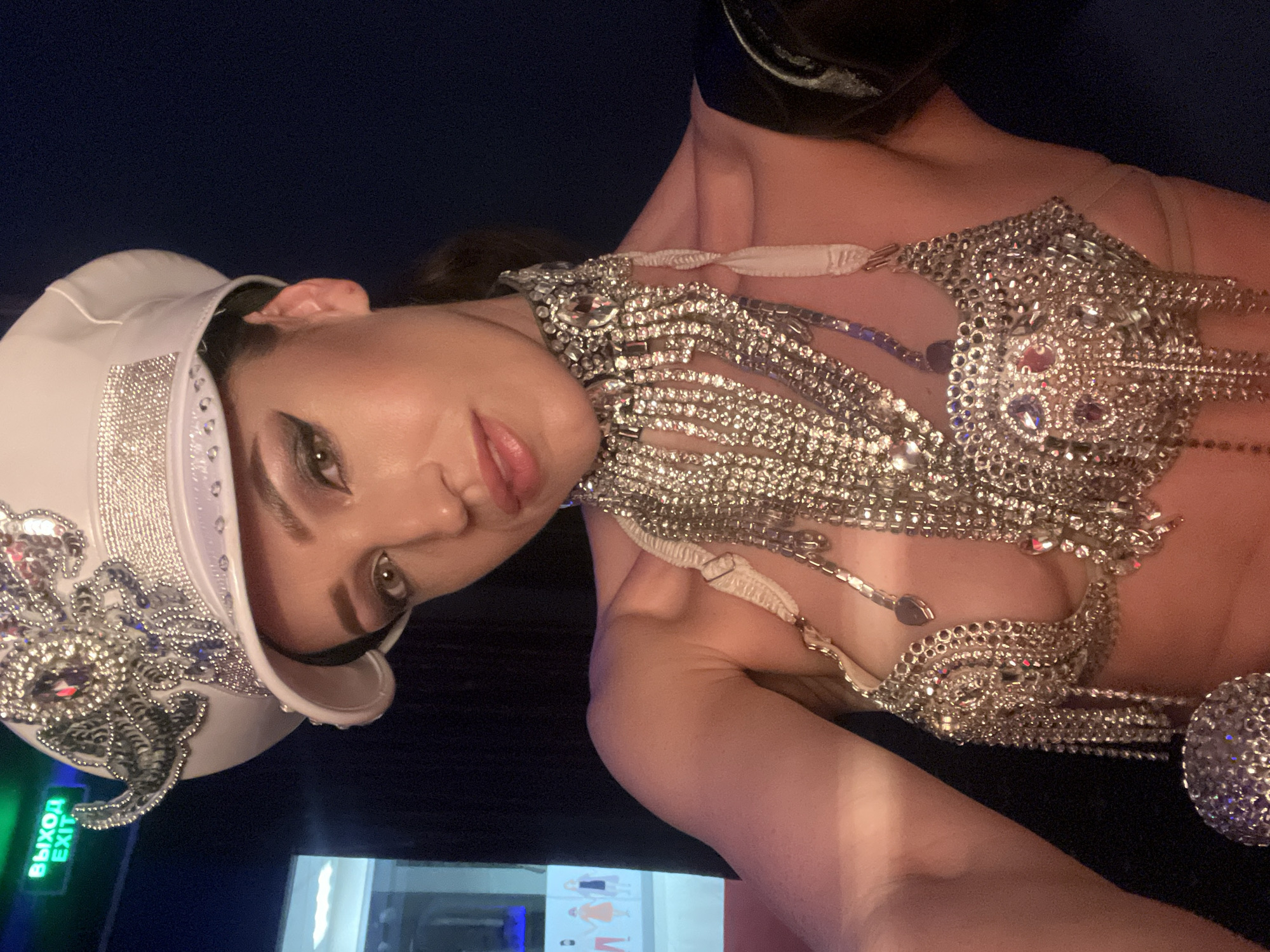 An adult female, positioned in a close-up selfie, occupies the majority of the frame. Her head is adorned with a white military-style cap, heavily embellished with silver sequins and faceted clear and magenta gemstones, featuring a band of smaller rhinestones around the crown and brim. She wears a elaborate, silver-toned costume consisting of a wide choker-style necklace and a bralette, both encrusted with numerous clear and colored rhinestones and featuring cascading chains of rhinestones. A light-colored fabric strap is visible over her right shoulder. Her makeup includes prominent winged eyeliner and glossed lips. She looks directly into the camera. The background is dimly lit, primarily dark blue or black. In the upper left quadrant, a green illuminated "ВЫХОД EXIT" sign is visible, indicating a Russian-speaking location. Below the sign, a blurred display or reflection shows stylized, simplified figures of people against a lighter background. The scene is located indoors, possibly a performance venue or club, within the city of Kazan, Russia.