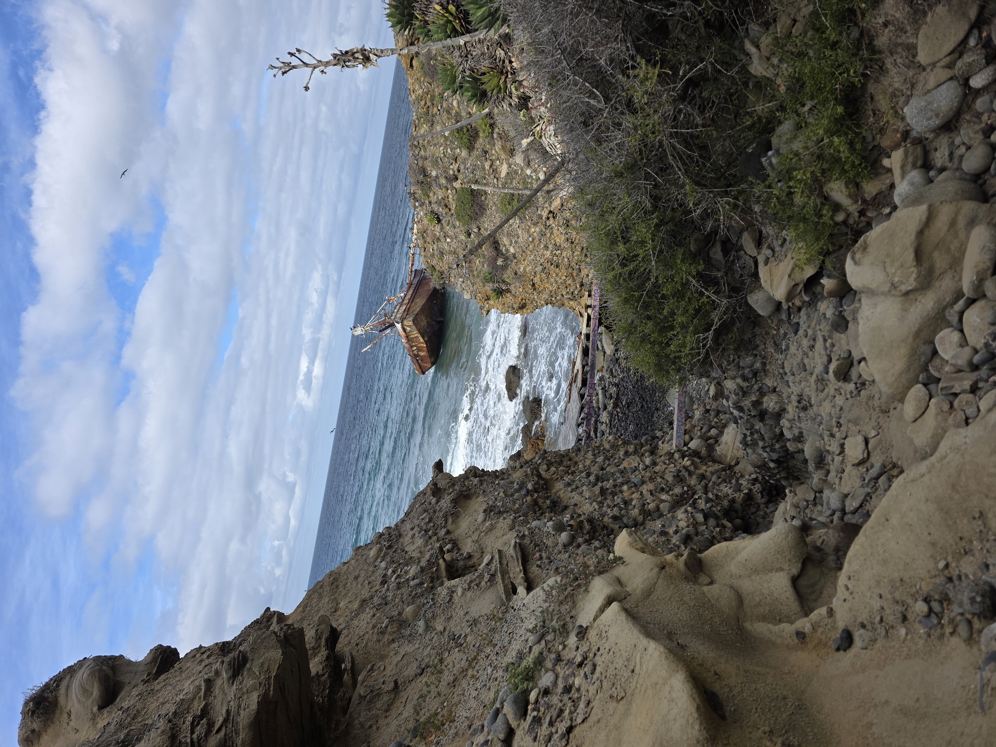 A medium, oblique-angle outdoor shot depicts a shipwreck located in Ensenada, Mexico. The scene is framed by coastal cliffs in the foreground. The rusted remains of the ship rest in the ocean's waves, positioned between two rocky cliffs. The ocean is a mix of dark blue and light blue tones. The sky above is partially covered with puffy white clouds, with patches of blue visible. A bird is seen flying in the upper left corner of the image. The cliffs in the foreground consist of rocks, pebbles, and sparse vegetation. The cliffs are shades of brown, gray, and tan.