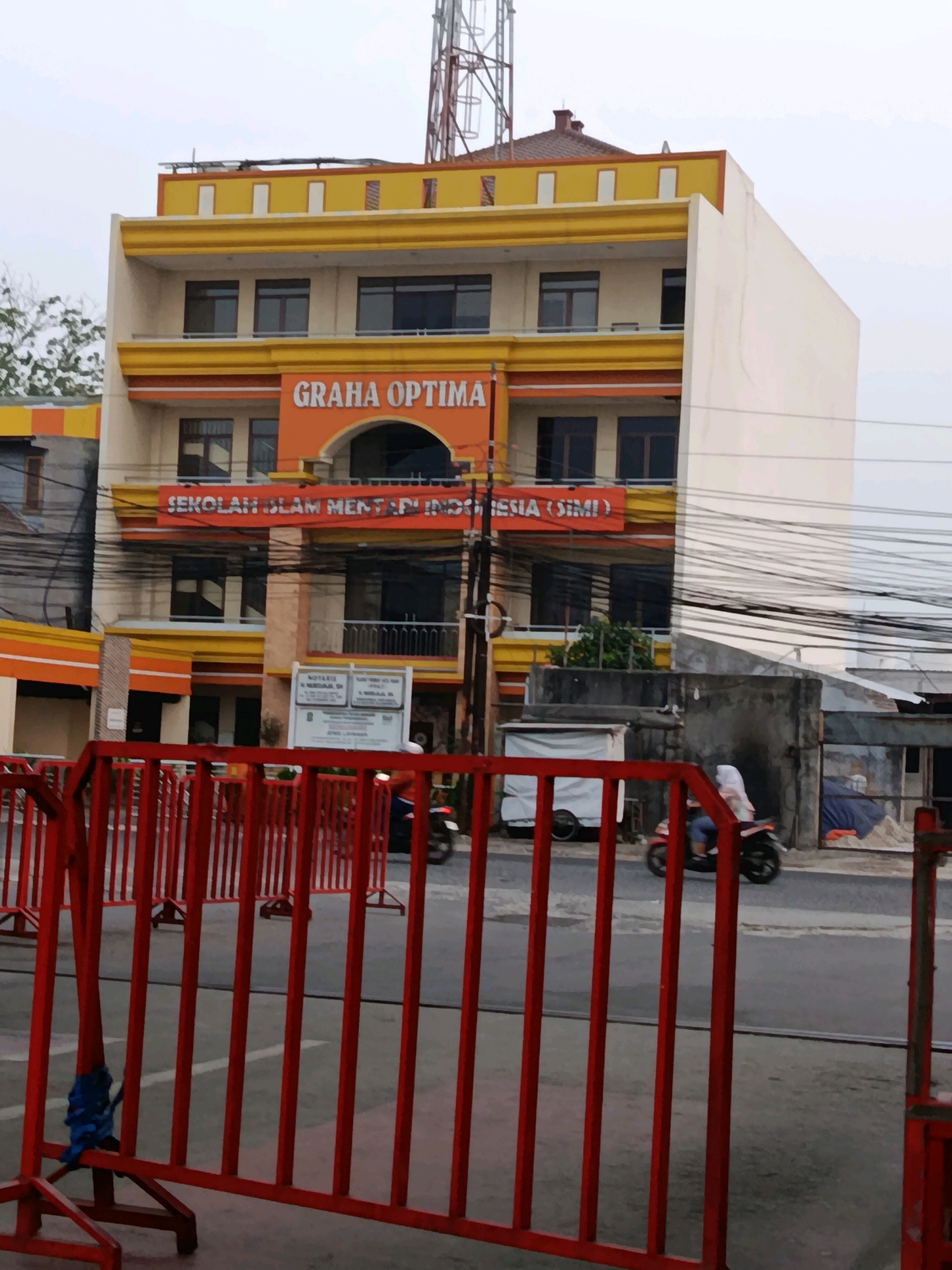 The image depicts the facade of a multi-story building with the prominent signage "GRAHA OPTIMA" and "SEKOLAH ISLAM MENTARI INDONESIA (SIMI)" in orange lettering on an orange background. The building is painted in yellow and orange, with cream-colored sections. Multiple windows with dark frames are visible on the upper floors. A telecommunications tower is partially visible at the top right of the frame. In the foreground, red metal barricades are set up, with a blue tie securing one of the barricade poles. A motorcycle with a rider wearing a white headscarf and blue clothing is visible on the road behind the barricades. The environment appears to be an urban setting, with power lines crisscrossing the scene and other structures visible in the background. Based on the signage, the location is identified as a school in Bekasi, Indonesia.