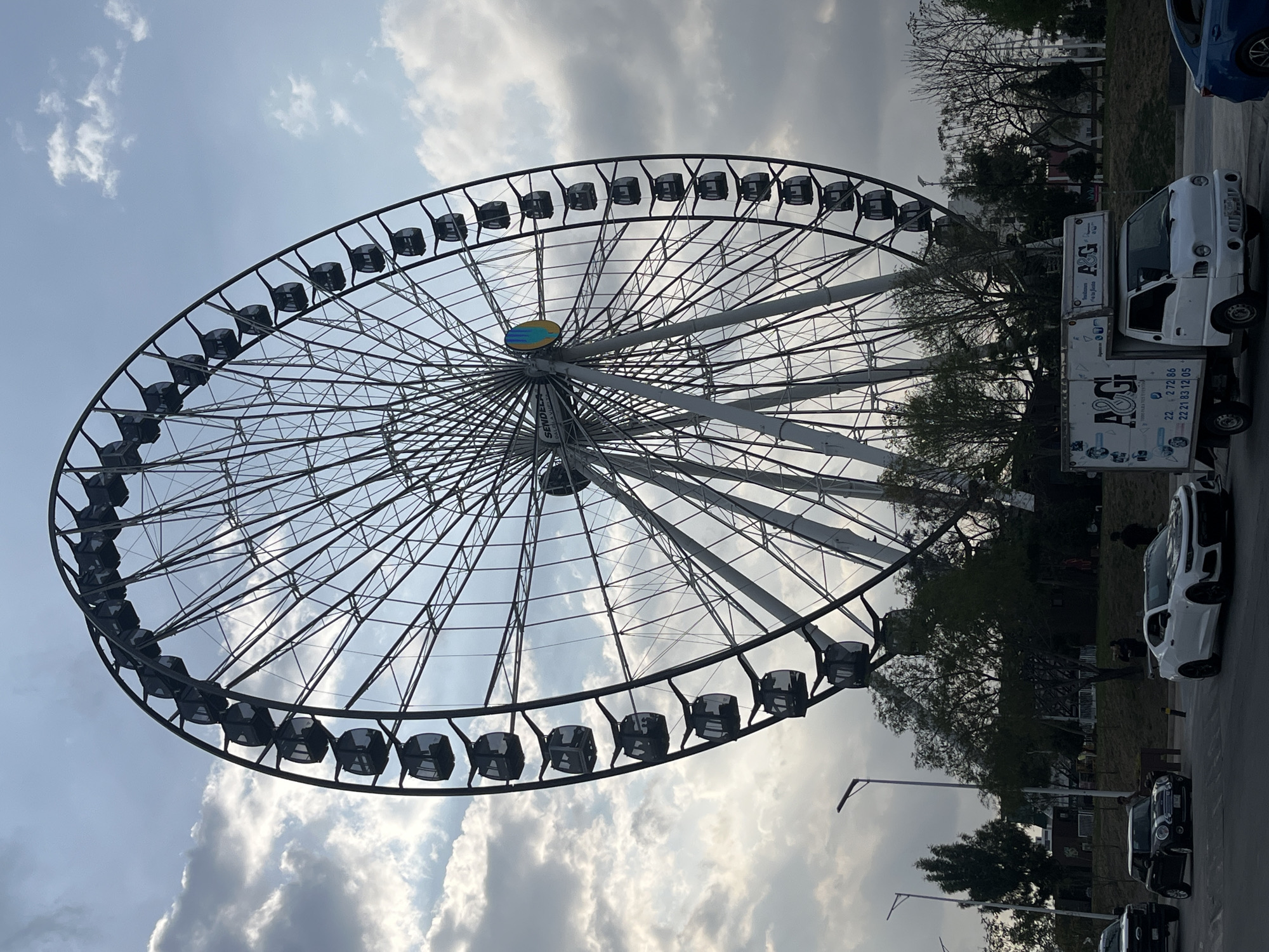 This image shows a large Ferris wheel with black gondolas against a cloudy sky. The wheel has a blue and yellow design at its center. To the right of the Ferris wheel, there are trees and parked vehicles, including a white truck with "A&GI" printed on its side. A road stretches out in the foreground with streetlights. The location appears to be in a city, potentially Unknown, Unknown, as indicated by the presence of urban infrastructure and vehicles.