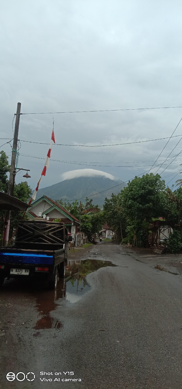 This image depicts a street scene in Pesawahan, Indonesia, under an overcast sky. A dark gray asphalt road, featuring multiple puddles reflecting the sky and surrounding elements, occupies the lower and central portions of the frame, receding into the distance.
In the immediate foreground on the left, a black Suzuki pickup truck is parked on the wet asphalt, facing away from the camera. Its rear is visible, showing a blue bumper, and a license plate (N 8325 NN, with a "02 27" tag). The truck bed features a wooden cage-like structure. To the left of the truck, a wooden utility pole with power lines and a hanging street lamp is visible. From a point above the pole, a vertical string of red and white banners hangs, possibly indicating Indonesian national colors.
In the midground, beyond the pickup truck and utility pole, various residential structures with tiled roofs are interspersed among dense green foliage and trees lining both sides of the road. Further down the road, a solitary individual on a dark motorbike is observed moving away from the camera.
The background is dominated by a large mountain, its peak partially obscured by a distinctive, elongated lenticular cloud formation. The entire sky is a uniform, light grey, indicating an overcast condition. The wet ground and puddles suggest recent precipitation.