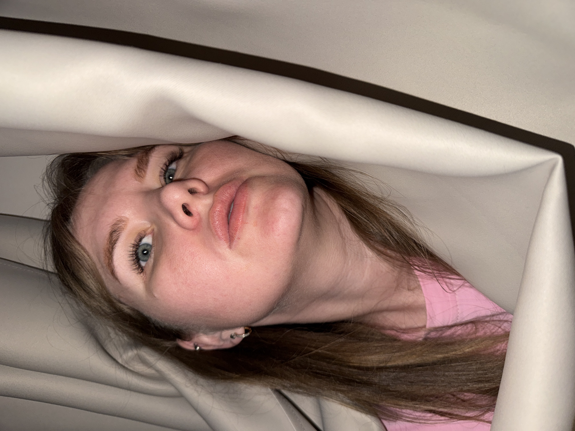 A close-up vertical shot in Gornyi (Dvurechenskoe S/P), Russia, depicts an adult female subject partially concealed by and looking through two light-colored, possibly beige, fabric panels resembling curtains. Her head is tilted slightly back, and her blue eyes are directed upwards and to the viewer's right. She has long, light brown hair, visible along her left shoulder and cascading over her right. She wears a pink, ribbed-knit garment. Her visible left ear features a small hoop earring in the lobe and a bar-shaped piercing in the upper cartilage. The fabric panels create distinct folds and shadows, framing her face and obscuring the background entirely.