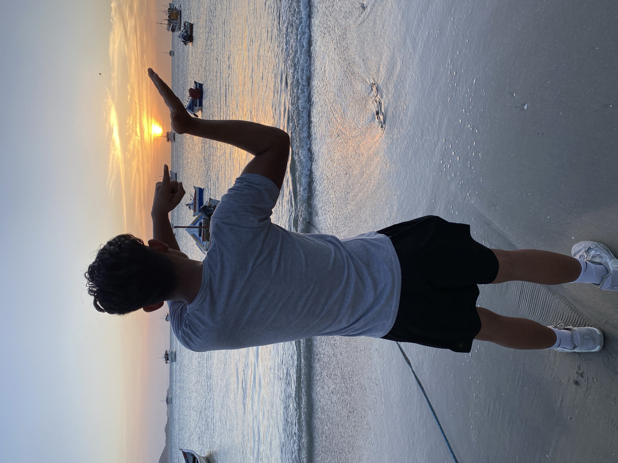 A person stands on a beach, facing away from the viewer, toward the ocean and a vibrant sunset. The individual is wearing a light gray t-shirt and dark shorts, with white shoes and socks. Their arms are raised and bent, with their fingers pointing in the general direction of the sunset. The beach appears to be sandy, with small waves gently lapping at the shore. In the background, several boats are visible on the water, silhouetted against the sunset. The overall lighting suggests it is either sunrise or sunset, with warm tones dominating the sky and reflecting off the water and sand. The location is Juan Griego, Venezuela.