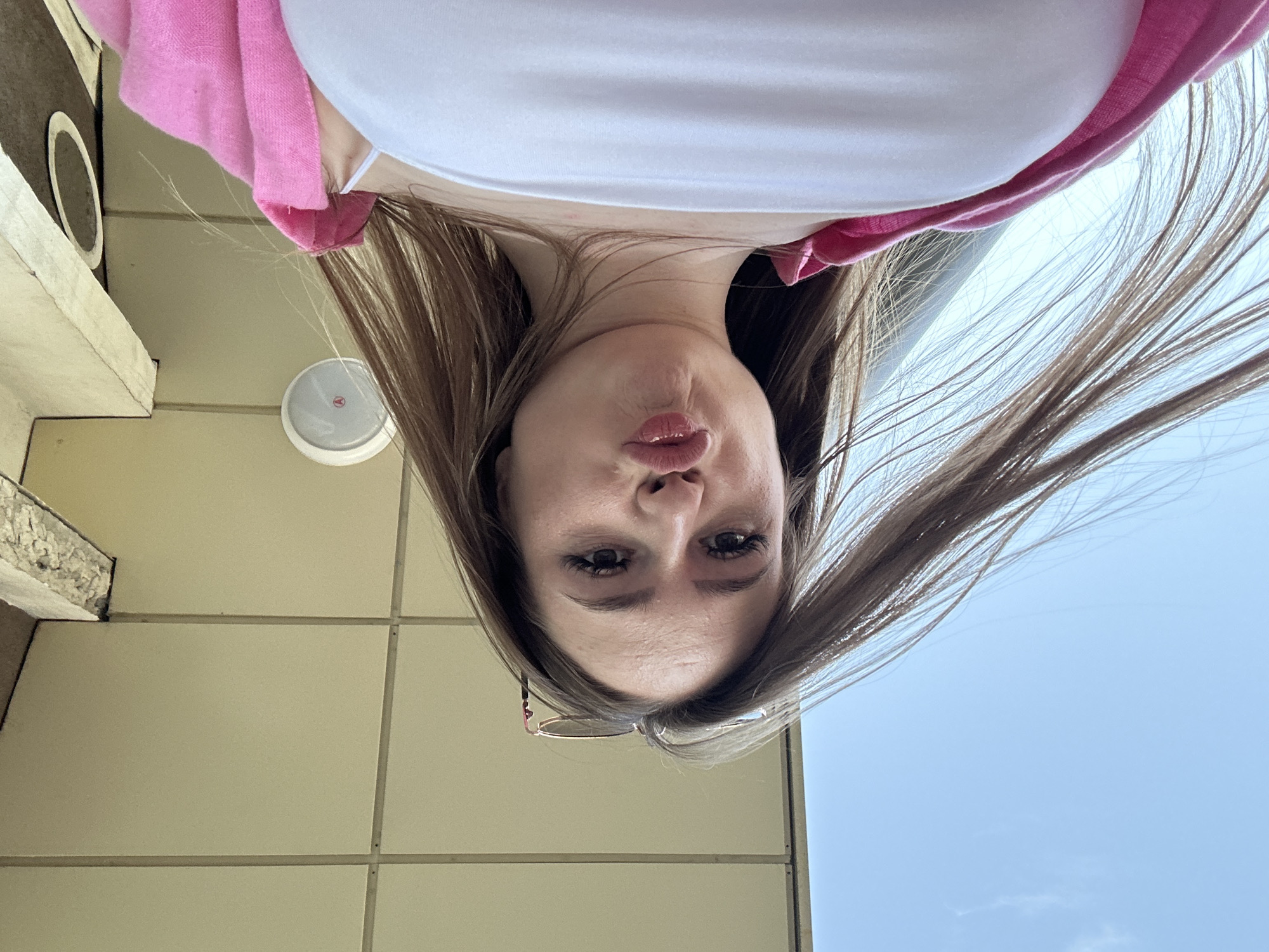 A young woman with long brown hair, wearing a white top and a pink cardigan, is the primary subject of this image. She is looking directly at the camera with her head tilted, and her lips are slightly puckered in a playful expression, possibly a kissy face or a pout. Her eyelashes and eyebrows are prominent, and a pair of glasses is visible resting on her head or hair near her temple.
The photograph is taken from a very low angle, looking upwards, which causes the woman to appear as if she is looking down from above. The background consists of a light-colored building wall, which appears to be tiled or panelled, with visible grout lines in some areas. Affixed to this wall is a circular white object featuring a red triangular logo, possibly with the letter "A" inside it. To the left, a concrete or stone architectural ledge is partially visible. The upper right portion of the image (when mentally oriented upright) shows a clear, light blue sky, indicating daytime and fair weather. Her hair appears to be flowing, consistent with the image's perspective and potential movement.
The overall scene suggests an outdoor urban or suburban setting, likely next to a building. There is no other discernible text apart from the logo on the circular object. The image conveys a playful and expressive mood from the woman.