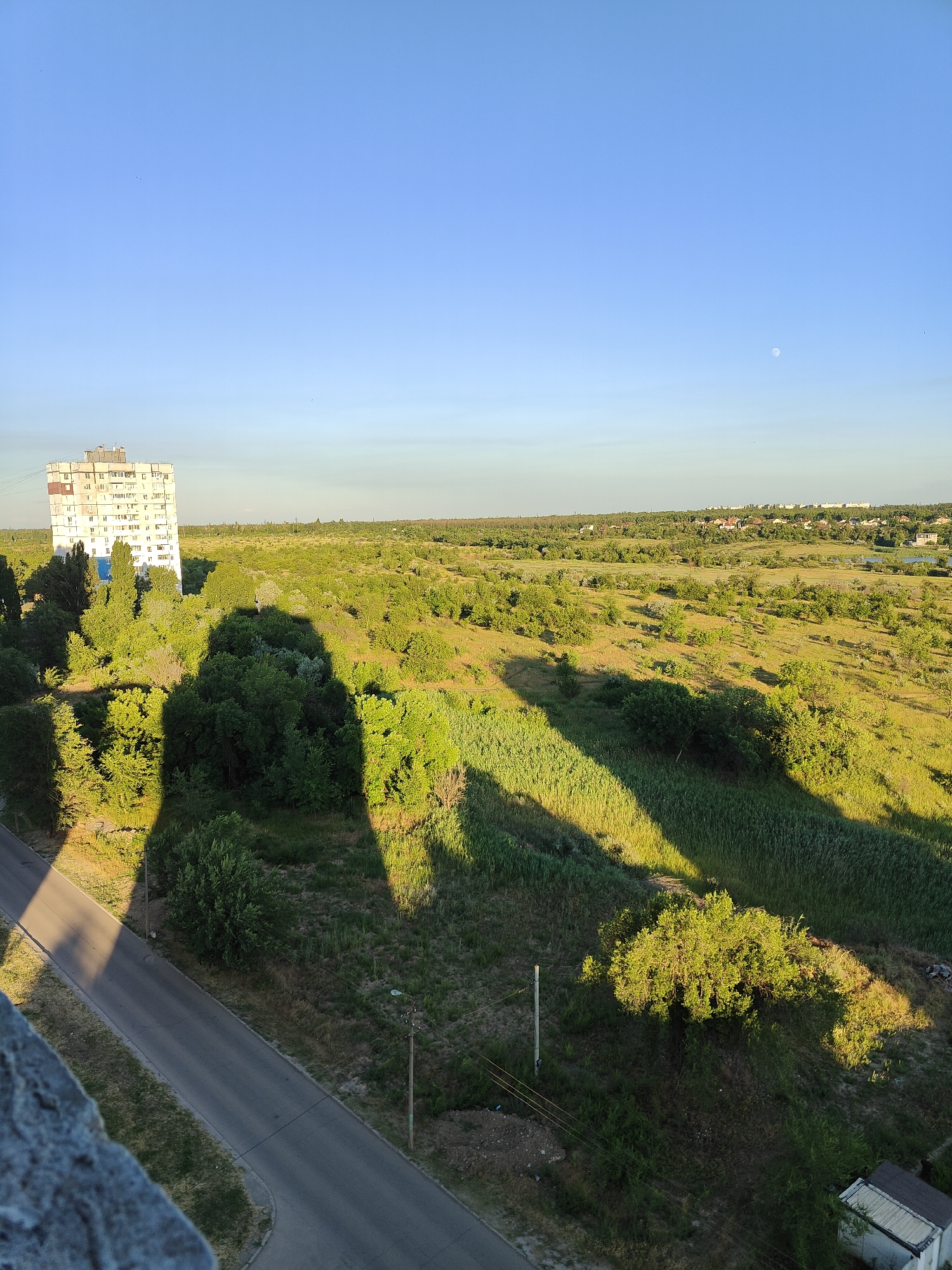 This image depicts an aerial view of a rural landscape bathed in the warm light of late afternoon or early morning. The dominant feature is a vast expanse of green, undulating terrain covered in dense vegetation, including trees, bushes, and tall grasses. Long, dramatic shadows are cast across the landscape, indicating the low angle of the sun.
In the upper left quadrant, a tall, multi-story apartment building stands out against the natural backdrop. It appears to be an older structure, with a somewhat utilitarian design. A paved road snakes its way through the lower left of the frame, bordered by the grassy embankment.
In the distance, a hazy line of trees and the rooftops of houses suggest a small settlement or village. The sky is a clear, pale blue, with a faint, barely visible moon high up on the right. The overall impression is one of quietude and a peaceful natural environment contrasted with man-made structures. The lighting suggests a time of day when the sun is low, casting long shadows, characteristic of either sunrise or sunset. The weather appears clear and calm. The location context of Kryvyi Rih, Ukraine, places this scene in a specific geographical region, likely characterized by such landscapes. There is no visible text in the image.