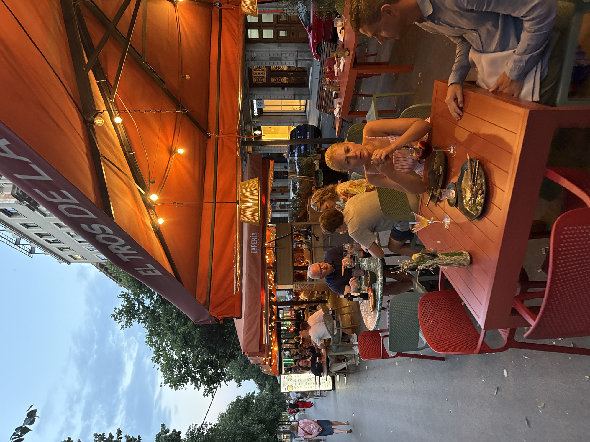 The image depicts an outdoor dining area of a restaurant named "EL TROS DE LA" which appears to be a "TAPERIA CATALANA" in Barcelona, Spain, judging by the name and the style of the establishment. The scene is set in the late afternoon or early evening, with warm lighting from overhead lamps and string lights illuminating the orange awnings. Several tables are occupied by patrons enjoying food and drinks.
In the foreground, a woman with blonde hair is seated at a red table, looking towards the camera with her hands clasped. Beside her, a man in a light blue shirt is seated and looking to his right, with plates and drinks on the table. A vase with dried flowers sits on the table.
In the middle ground, several other tables are visible with groups of people dining. One table features a man with glasses and a blue shirt, holding cutlery. Further back, more tables and patrons are discernible under the awnings, with some tables having patterned tabletops and colorful chairs. A menu board is displayed outside one of the restaurant sections.
The background shows a tree-lined sidewalk with people walking. Beyond the restaurant, buildings with multiple stories are visible, including a store with "STARBUCKS" signage and another establishment with ornate doorways. Cars are parked along the street and in the background. The overall environment suggests a lively urban setting in Barcelona.