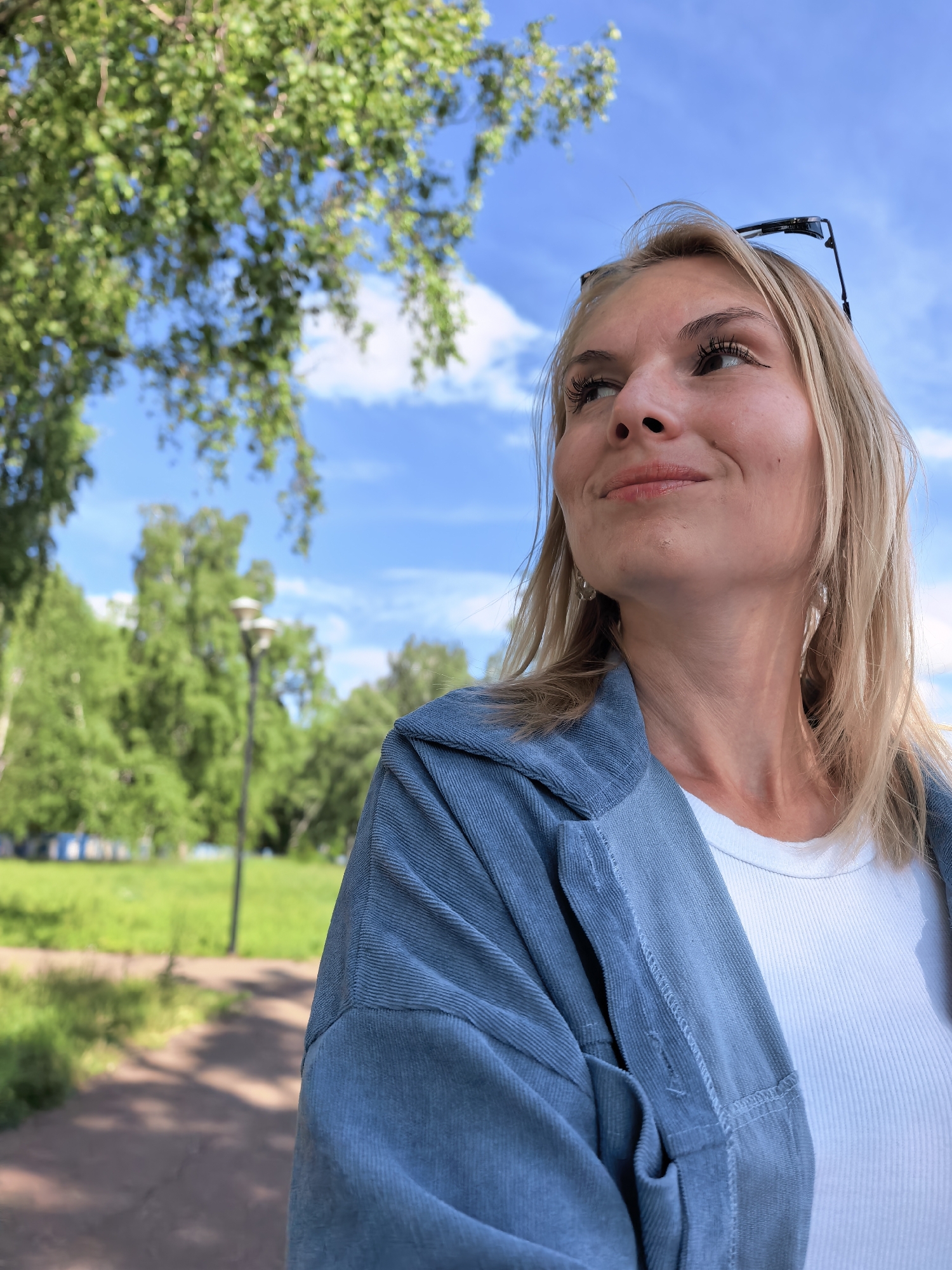 The image depicts an adult blonde female outdoors in a park environment, attributed to being in Sterlitamak, Russia, on a clear, sunny day. The woman is captured from a low angle, looking upwards and to her right with a slight smile. She wears a white ribbed top and an unbuttoned blue-grey corduroy-style jacket. Black-framed sunglasses are perched on her head, and she has silver-toned, elongated teardrop-shaped earrings. The background features a bright blue sky with scattered white clouds, out-of-focus green deciduous trees, an unpaved pathway in the lower left, and a tall lamppost in the mid-ground. Distant, blurred blue and white structures are visible amidst the trees.