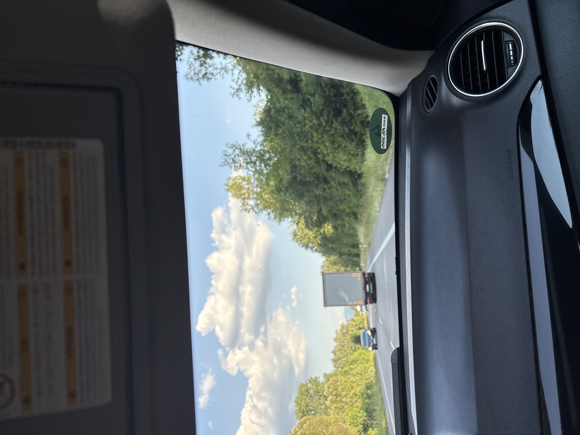 The image is taken from the interior of a car, looking out through the windshield. The view is of a highway with trees lining the sides. A gray truck is in the right lane, and a dark car is in the left lane, both moving away from the camera. The sky is blue with white clouds. The dashboard of the car is visible in the foreground, with an "AIRBAG" label and an air vent. A circular green sticker with white lettering is visible on the right side of the windshield. This sticker has the text "BAD VÖCK" in white, which is an abbreviation for Bad Vöcklin, a town in Germany. The image is likely taken in the city of Iffezheim, Germany, given the context of the sticker.