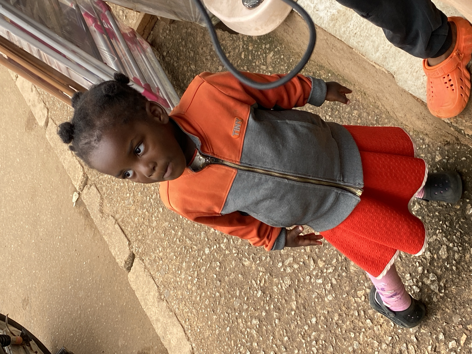 In the city of Bukuru, Nigeria, a young Black girl stands on a gritty, light brown dirt and gravel path, looking upwards. She is wearing a gray and orange zip-up jacket with the word "TINY" embroidered in orange on the left breast. Beneath the jacket, she wears a bright red, textured skirt. Her legs are clad in pink socks with a floral pattern, and she has on black slip-on shoes. The girl's hair is styled in small buns. To her right, a partially visible person wearing black pants and an orange Croc shoe stands on a raised concrete curb. Behind the girl, several metal poles covered in clear plastic are leaning against a structure. The scene appears to be outdoors, possibly near a market or street vendors.