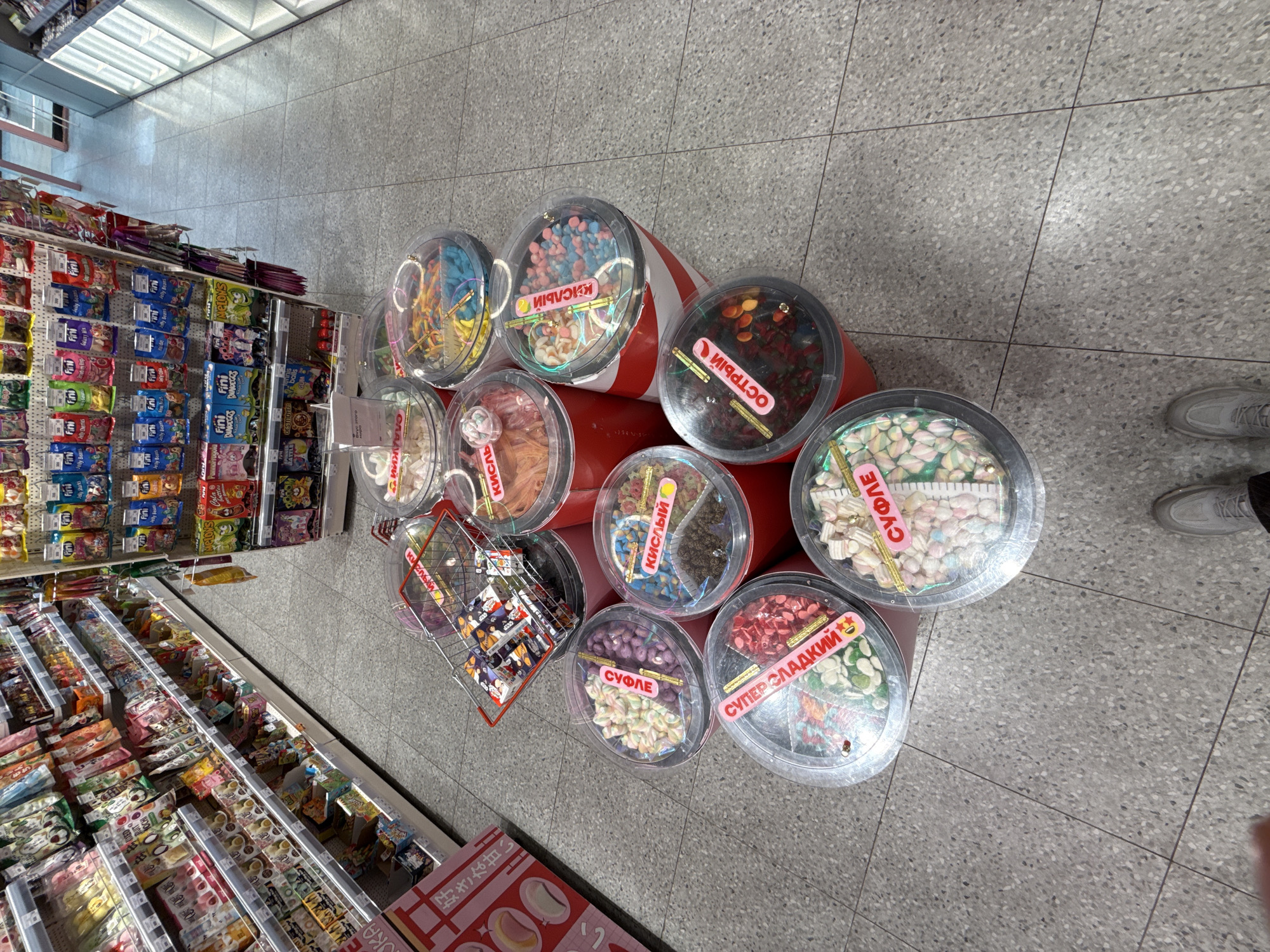 The scene appears to be inside a store, potentially a grocery or convenience store, located in Moscow, Russia. Aisles of shelves stocked with various packaged candies and snacks are visible. At the end of the aisle are large, clear containers filled with different types of bulk candies. The containers are labeled in Russian, and feature categories such as "sour," "sweet," "souffle," and "spicy." A small shopping basket containing items is placed near the containers. The floor is tiled in a gray and white pattern. The image is taken from an elevated, slightly angled perspective. A person is visible in the bottom right corner with gray sneakers.