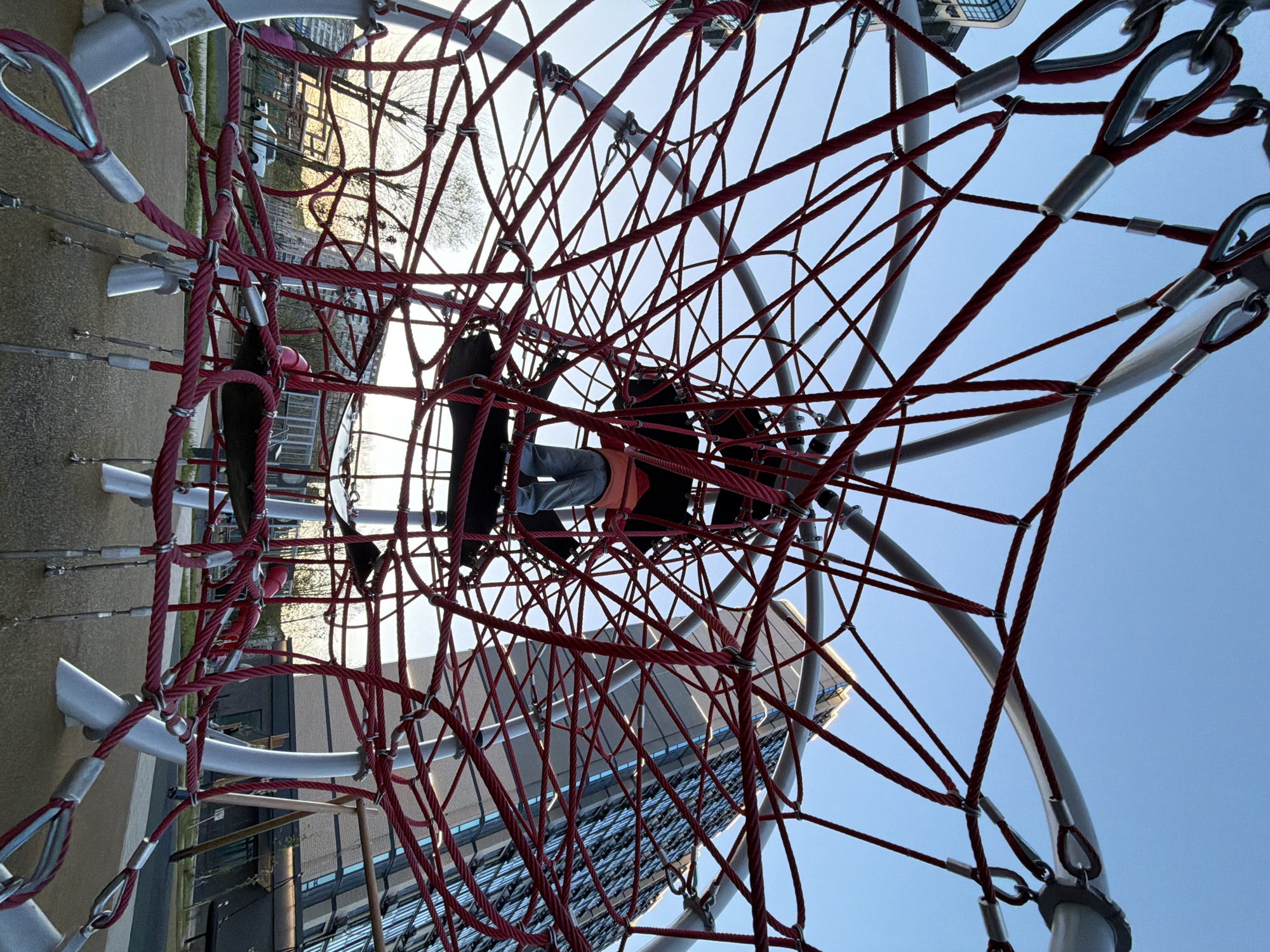 This image captures a vibrant, complex red rope climbing structure, likely part of a modern playground, set against a backdrop of urban architecture and a clear sky. The image is oriented sideways, with the sky appearing on the right and the ground and buildings on the left, suggesting a rotation from its original capture.
The central focus is a large, intricate net-like structure made of thick, bright red ropes. These ropes are interconnected at various points by silver metal clamps and secured to sturdy, light grey/silver metal poles with robust carabiner-like fittings and thimbles, indicating durable construction. Several individuals, likely children or young adults, are visible within the rope net. One person, wearing a grey shirt and what appears to be an orange safety harness or vest, is climbing or resting in the lower-middle section of the net. Another person, mostly obscured by the ropes, is positioned to the right of the first, wearing dark clothing. Their limbs are extended, gripping the ropes, indicating active play.
In the background, modern buildings with reflective glass facades rise upwards, suggesting an urban or suburban setting. Bare trees, possibly indicating autumn or winter, are also visible, contrasting with the contemporary architecture. The sky is a bright, clear blue, suggesting good weather conditions. The lighting appears to be either late afternoon or early morning, with the sun casting a warm glow on the distant buildings and filtering through the ropes. The ground immediately surrounding the structure is light-colored, possibly a soft-fall surface like sand or rubber. There is no visible text in the image.