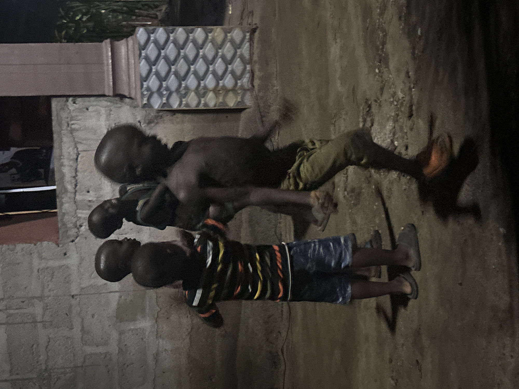 Three males are engaged in wrestling on a paved surface. The location appears to be outdoors near a building. One man is shirtless, wearing pants and shoes. One child is wearing a long-sleeved shirt and shorts, also wearing shoes. One child is wearing a t-shirt and jeans.