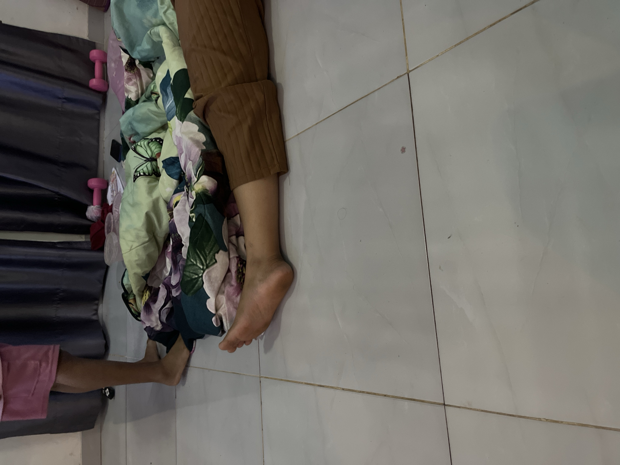 The image depicts a domestic indoor scene, likely a bedroom or living area, with a tiled floor. In the foreground, a person's legs are visible, one outstretched and resting on a patterned blanket featuring flowers and butterflies. The person is wearing brown, ribbed pants. To the left, another person's legs are also visible, wearing a pink skirt and standing barefoot. In the background, dark curtains hang behind a pile of blankets and what appear to be pink dumbbells. The overall impression is one of casual relaxation or a staged setting. The location is not specified in the image.