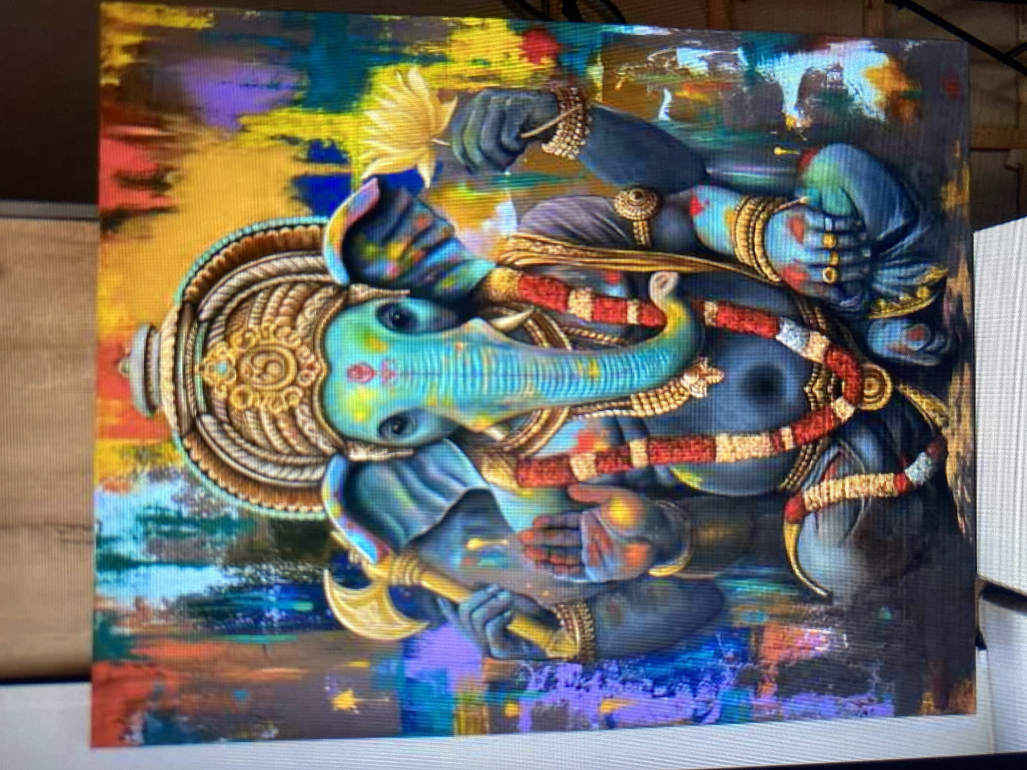 An abstract painting of Ganesha, the Hindu deity, is displayed on a canvas. Ganesha is depicted with a blue elephantine head and a human-like torso, seated cross-legged. He has four arms, with the upper right arm holding a golden axe, and the upper left arm holding a yellow lotus flower. His lower right hand is raised in a blessing gesture, and his lower left hand rests on his knee. He is adorned with a golden headdress, a red and gold garland, and elaborate golden bracelets and armbands. The background of the painting is a vibrant, abstract composition of vertical strokes in various colors, including red, orange, yellow, blue, green, and purple, creating a dynamic and energetic effect. The overall style of the painting is colorful and somewhat impressionistic, with visible brushstrokes and textured application of paint.