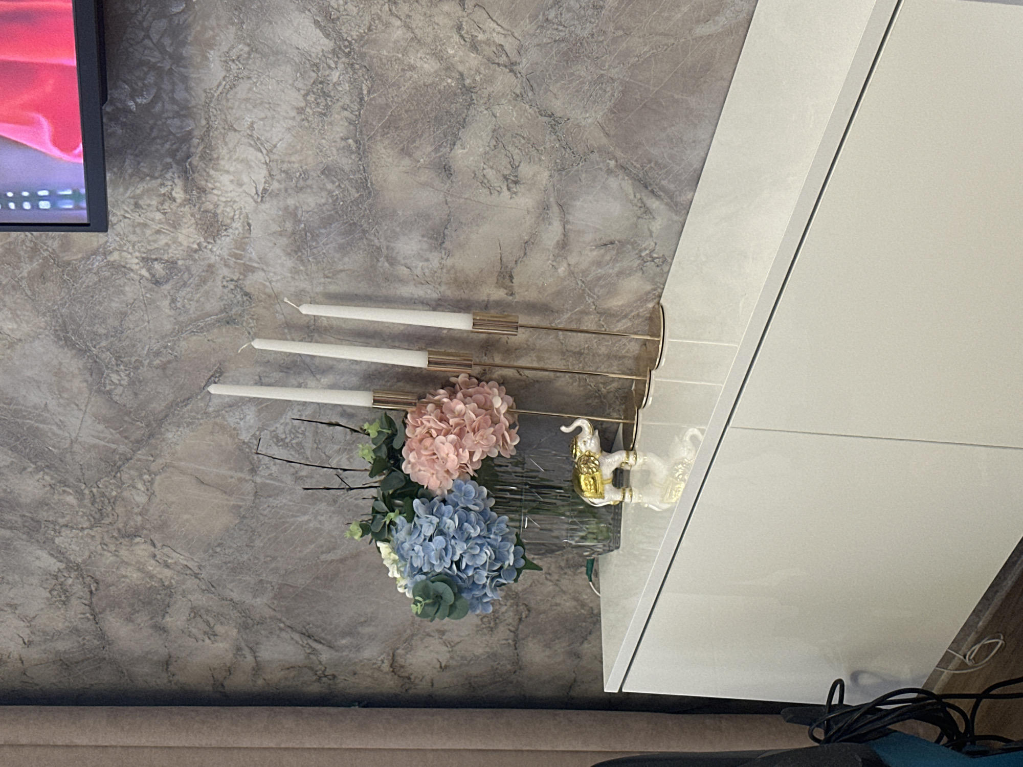 The image displays a modern interior scene, likely a living room or dining area, with a focus on decorative elements. The background features a wall with a marble-like texture in shades of grey, suggesting a sophisticated aesthetic. On the wall, a three-arm candelabra holds unlit white candles. To the right of the candelabra is a decorative glass vase containing a bouquet of artificial flowers, predominantly in shades of pink and blue, with greenery. Next to the vase, a small, white decorative elephant with gold accents is placed.
In the upper left corner, a portion of a television screen is visible, displaying a bright red and pink abstract image. The furniture in the foreground is sleek and contemporary, with a white, glossy cabinet or shelf unit occupying the right side of the frame. The lighting appears to be interior, possibly natural light supplemented by artificial sources, casting soft shadows. The overall mood is calm and refined, indicative of a well-decorated living space. The image is oriented sideways. No visible text is present in the media file. The location is specified as Abakan, Russia, but this context is not directly evident from visual cues within the image itself.