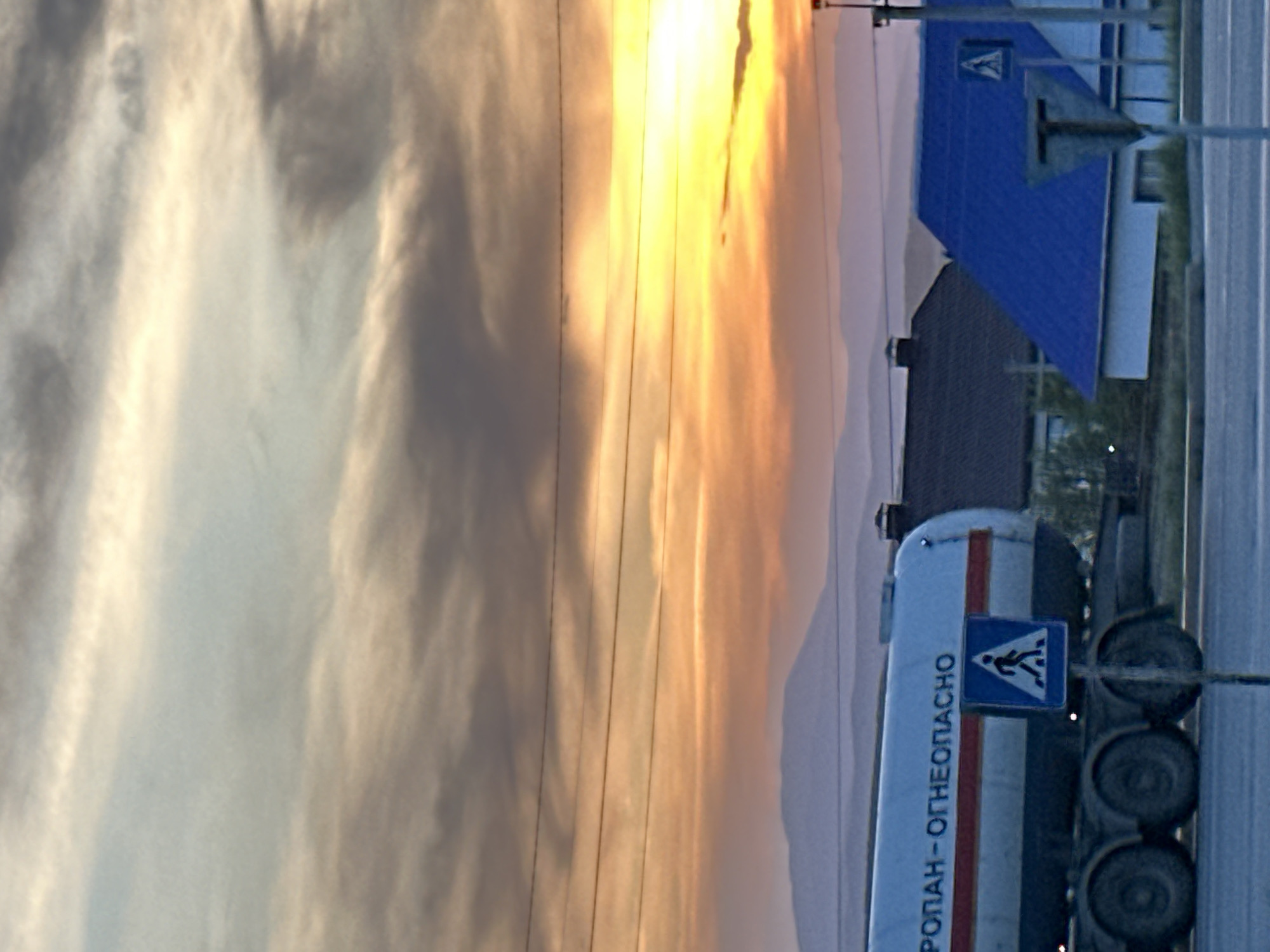 Here's a matter-of-fact description of the image:
The image is a landscape shot, possibly taken at dusk or dawn given the sky color. A tanker truck, with visible text in a Cyrillic script that includes the word "ОГНЕОПАСНО" (likely meaning "flammable" or "dangerous"), is positioned on the right side of the frame. A pedestrian crossing sign is affixed to the side of the tanker. A building with a blue roof and a prominent "A" symbol is in the background. Overhead power lines cross the upper center of the frame. A road appears to be on the right edge of the image. The location is labeled as Unknown, Unknown.