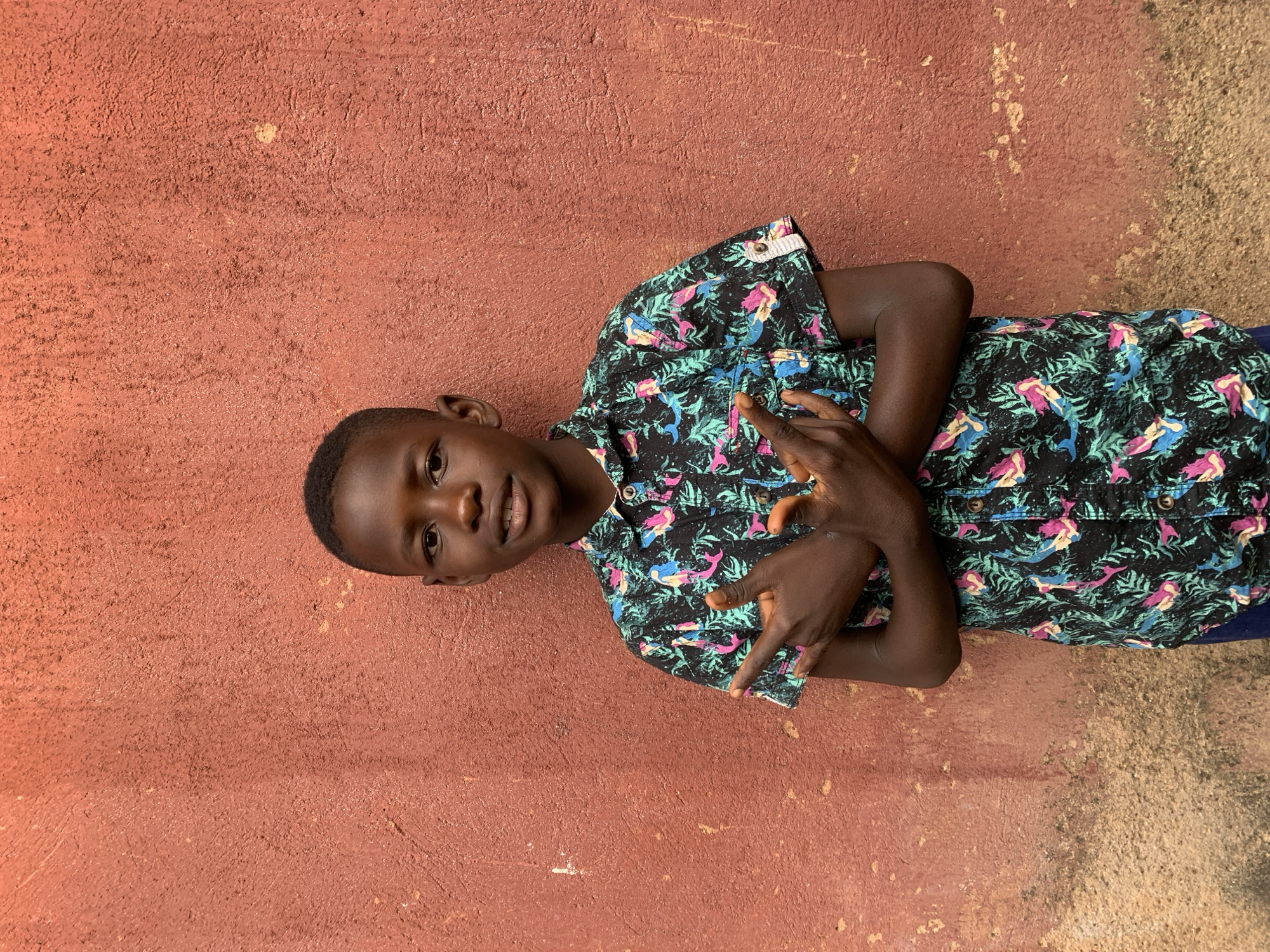 A young person with dark skin is standing near a reddish-brown wall. The individual is wearing a buttoned shirt with short sleeves and a pattern of green, pink, and blue. The person's hands are raised, fingers curled, forming a gesture. The background consists of a wall and ground, and is located somewhere in the city of Jos, Nigeria.