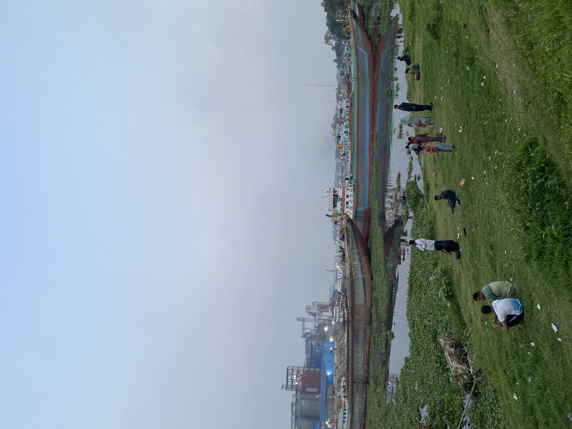 The image depicts a scene in Dhaka, Bangladesh, likely near the Buriganga River. Several large boats or ships are docked, with visible vegetation and structures associated with river transportation and industry. A group of people is present on a grassy bank alongside the river; their activities include sitting, standing, and possibly engaging in communal practices or social interactions. The sky is overcast, suggesting early morning or late afternoon lighting conditions. Debris is scattered on the grass.
