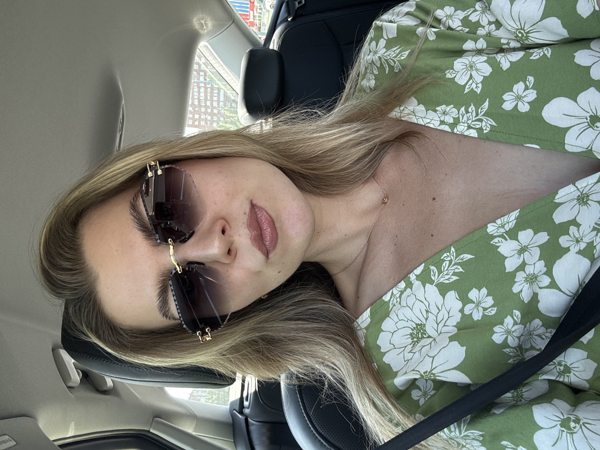A blonde woman with light skin and a floral print green top is seen from the chest up, sitting in the passenger seat of a car. She is wearing oversized, rimless sunglasses with a gold frame. Her mouth is slightly open, and she is looking towards the camera. She has a delicate gold necklace with a small pendant on her neck. The interior of the car is visible, with black leather seats and a glimpse of buildings and trees through the rear window. The lighting suggests it is daytime.