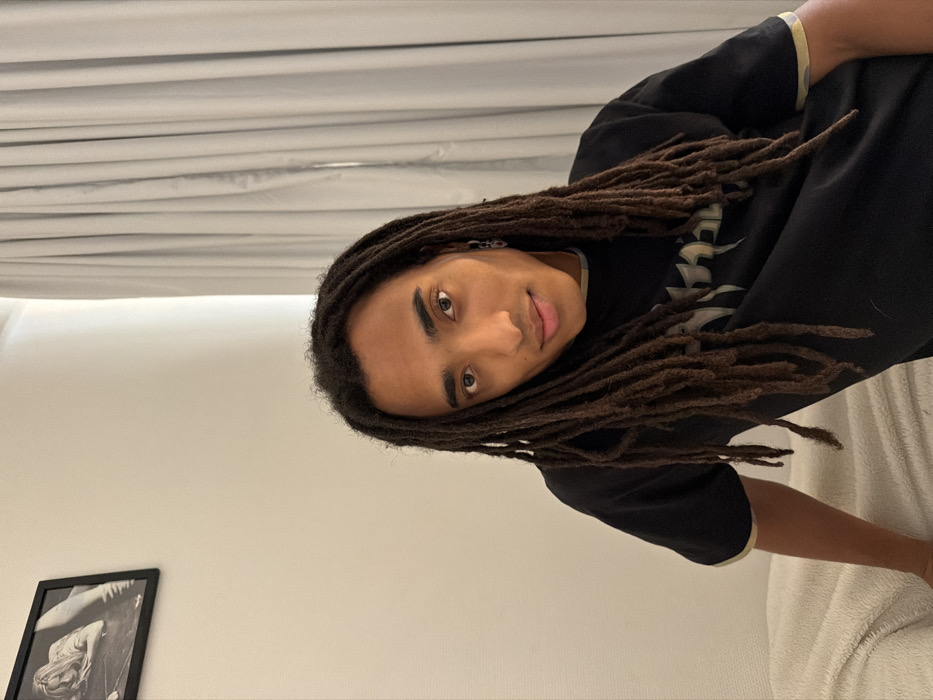 A young adult male with long, dark brown dreadlocks is depicted indoors, likely within the city of Paris, France. He wears a black short-sleeved t-shirt featuring a light-colored trim and a partially obscured graphic on the chest. A dark, possibly linked, earring is visible on his left ear. The individual is positioned with a slight head tilt, making direct eye contact with the viewer, and exhibits a subtle facial expression suggestive of a smirk or slight smile. His left arm rests upon a light-colored, textured textile, possibly a blanket or bedding. The immediate background to his left consists of a plain, light-colored wall, upon which a framed black-and-white photograph of a reclining figure is mounted in the upper left corner. To his right, light grey, drawn curtains cover a window or opening. The scene is evenly lit.