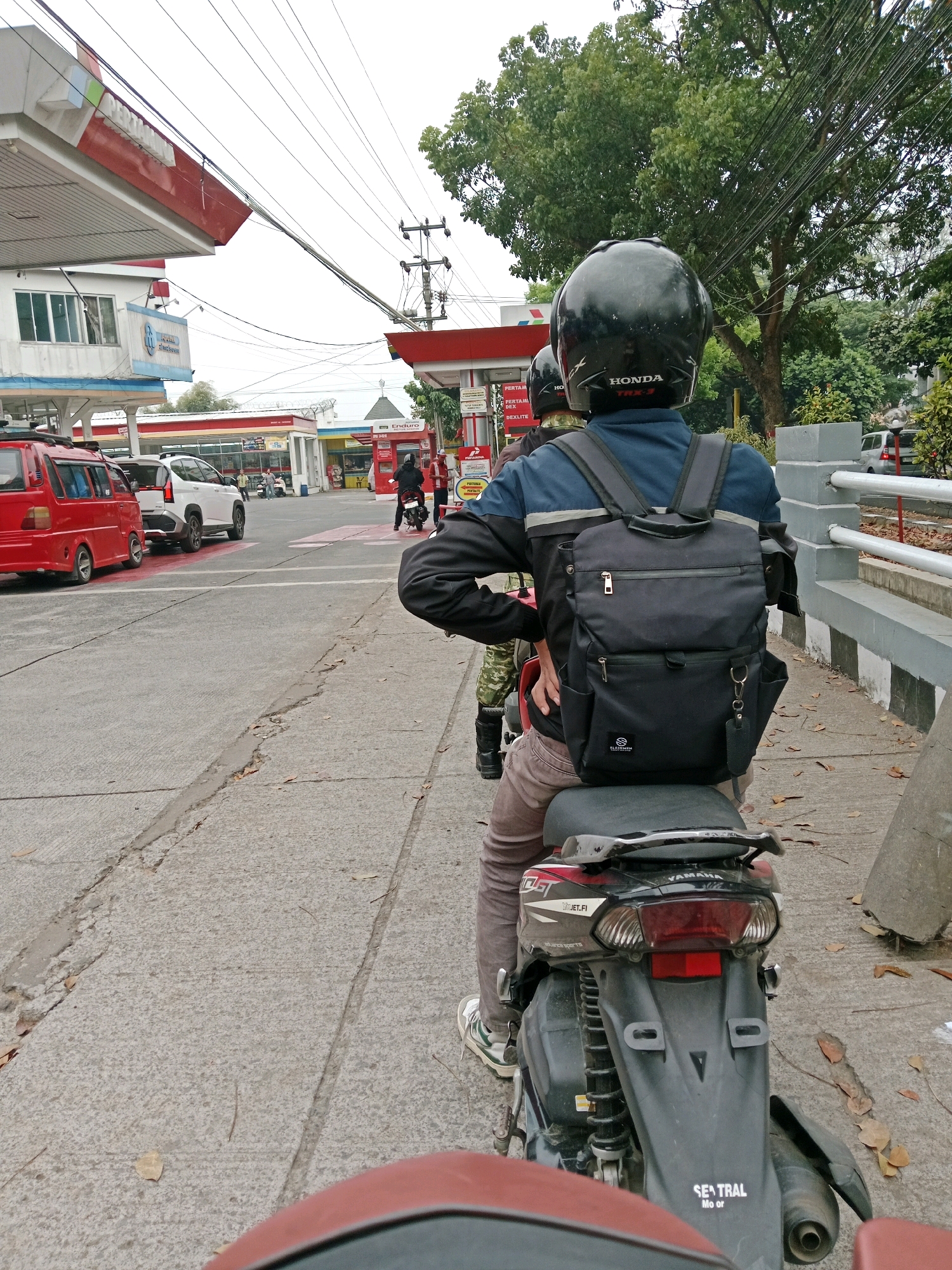 The image depicts a street scene at a Pertamina gas station in Muka, Indonesia. In the foreground, a person wearing a black helmet and a dark jacket with a backpack is seated on a motorcycle. The motorcycle is black with red accents and the brand "YAMAHA" is visible. The rider is facing away from the camera, looking towards the gas station. Behind this rider, another person on a motorcycle is partially visible. In the background, several vehicles are present, including a red van and a white SUV. The gas station itself has red and white structures with the Pertamina logo prominently displayed. There are also power lines and trees visible, indicating an outdoor urban environment. The ground is paved with concrete and shows some cracks and fallen leaves.