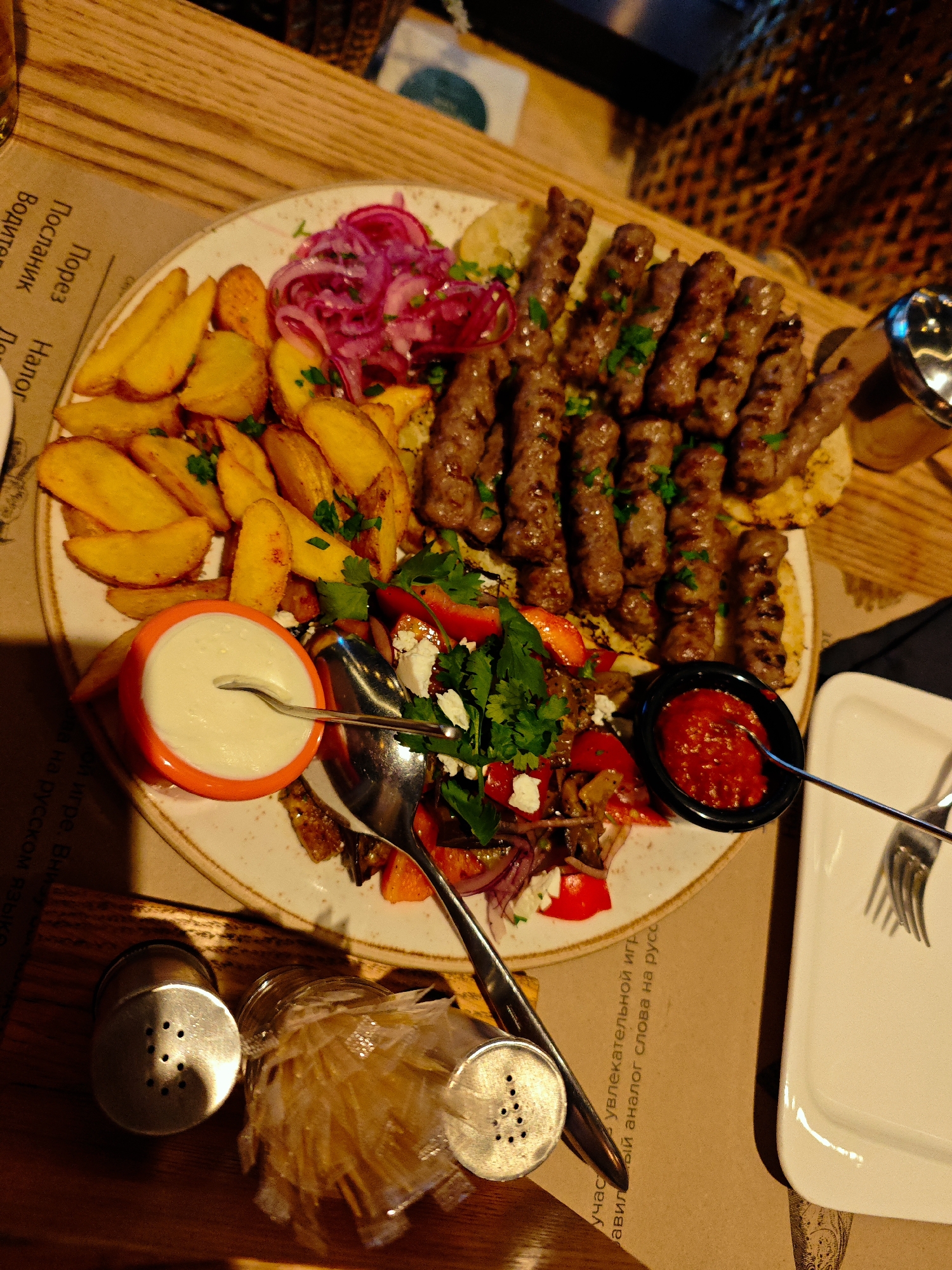 This image shows a close-up, overhead view of a platter of food on a wooden table. The platter contains several portions of grilled meat, likely kebabs, arranged in rows. Alongside the meat are golden potato wedges, a salad with chopped red peppers and feta cheese, and a dollop of white sauce in a small orange bowl. A small black bowl contains a red sauce, likely a relish or chili paste. Two metal serving spoons are visible, one in the white sauce and one in the red sauce. A fork rests on a white plate to the right of the platter. Two salt and pepper shakers and a glass jar filled with what appears to be dried pasta or noodles are also on the table. The background shows a woven basket and a wooden surface. The lighting suggests an indoor setting, possibly a restaurant. The text visible on the paper placemat is in Russian and appears to be part of a game or advertisement. The overall impression is of a hearty meal being served.