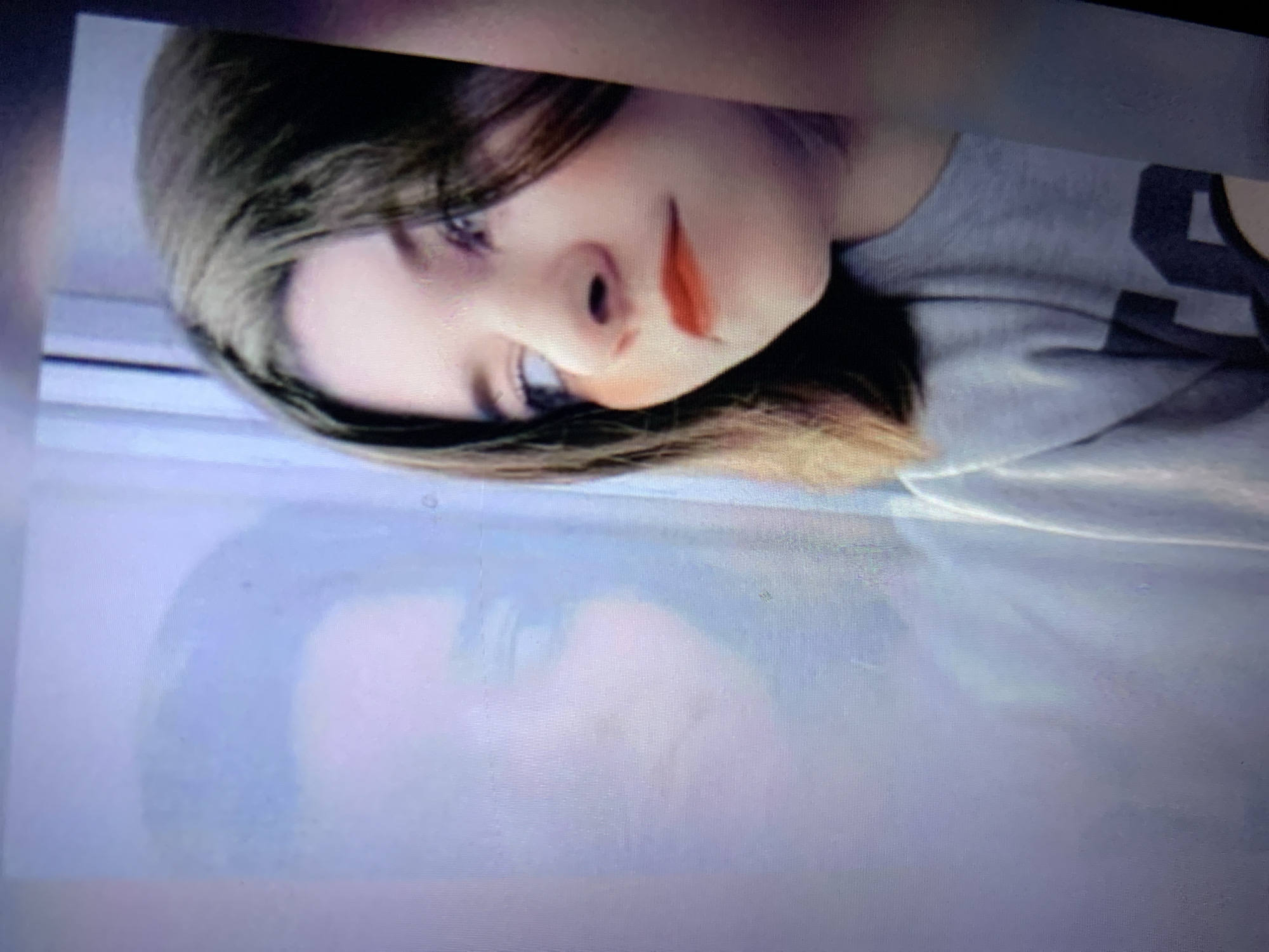 A young woman with dark hair and bright red lipstick looks to her left, her gaze directed slightly upwards. A faint reflection of her face is visible in a large window to her left. She is wearing a grey sweatshirt with dark lettering on the front. The background appears to be an indoor setting with a window and a vertical element, possibly a doorframe or part of the window structure, to the right of the woman. The lighting is soft, and the overall impression is one of quiet contemplation. The image does not contain any elements that would indicate a specific location within the city of Lymanka, Ukraine.