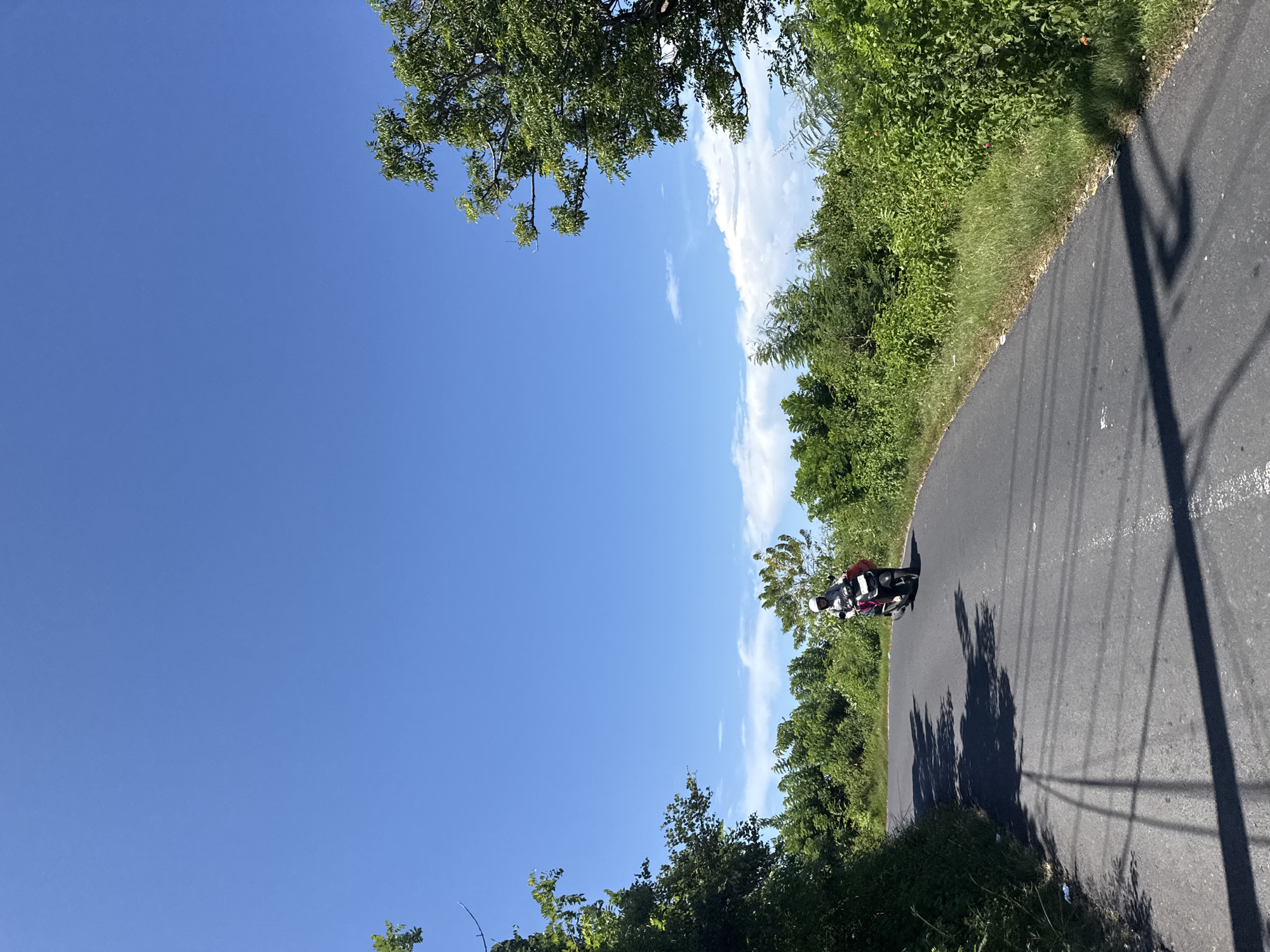 The image shows a paved road curving to the right. Lush green vegetation lines the right side of the road, and trees are visible at the top and bottom of the frame. On the road, a person wearing a helmet is riding a motorcycle. Shadows from overhead power lines fall across the road surface. The sky is predominantly blue with scattered clouds. The location is likely Batununggal, Indonesia, as specified in the prompt.
