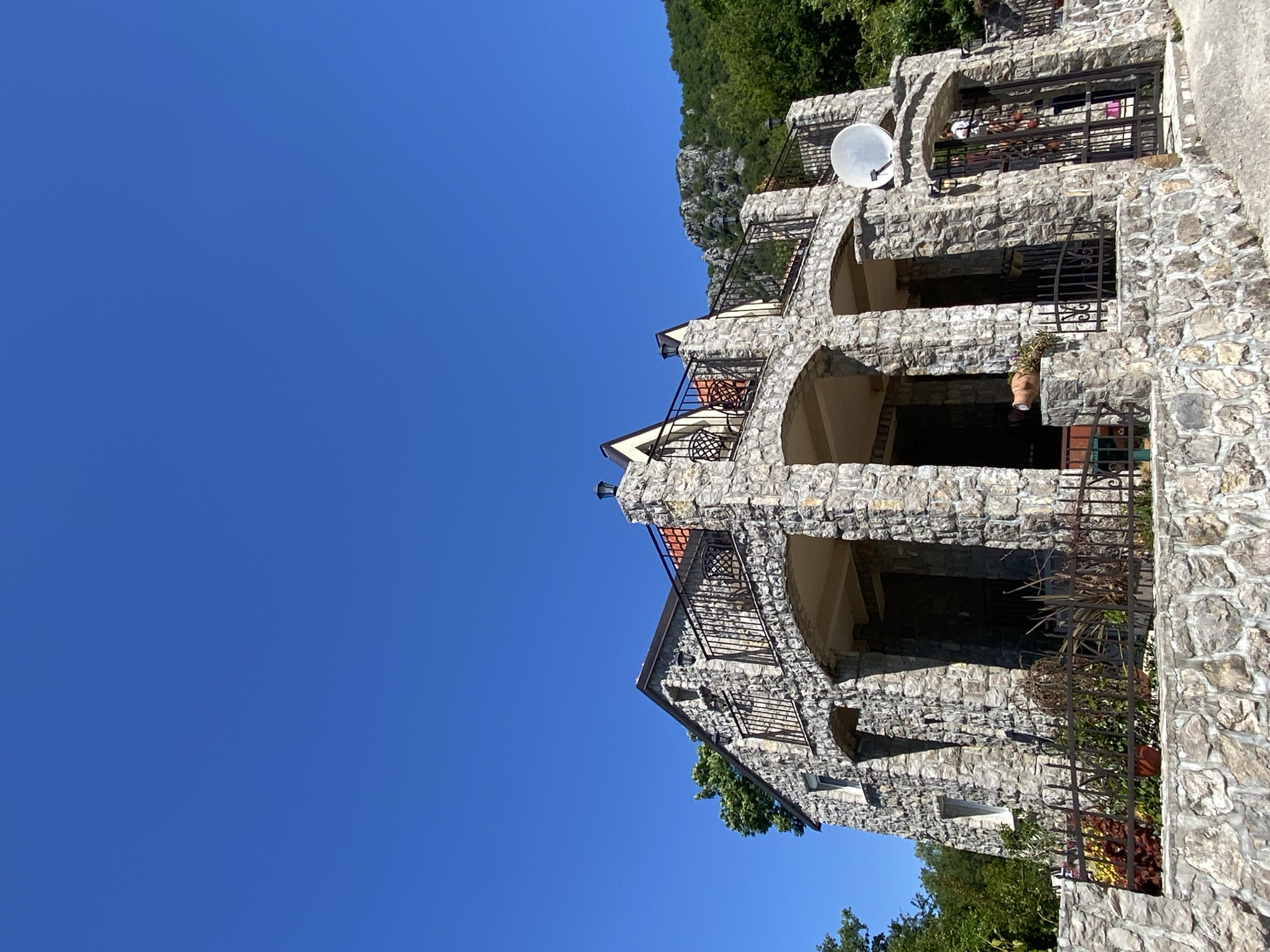 The image depicts a stone building in Sotonići, Montenegro, under a clear blue sky. The structure has multiple levels with arched openings and wrought iron railings. A satellite dish is mounted on the upper level. The building is constructed from roughly cut stones of varying sizes. The surrounding environment includes greenery and a hillside.