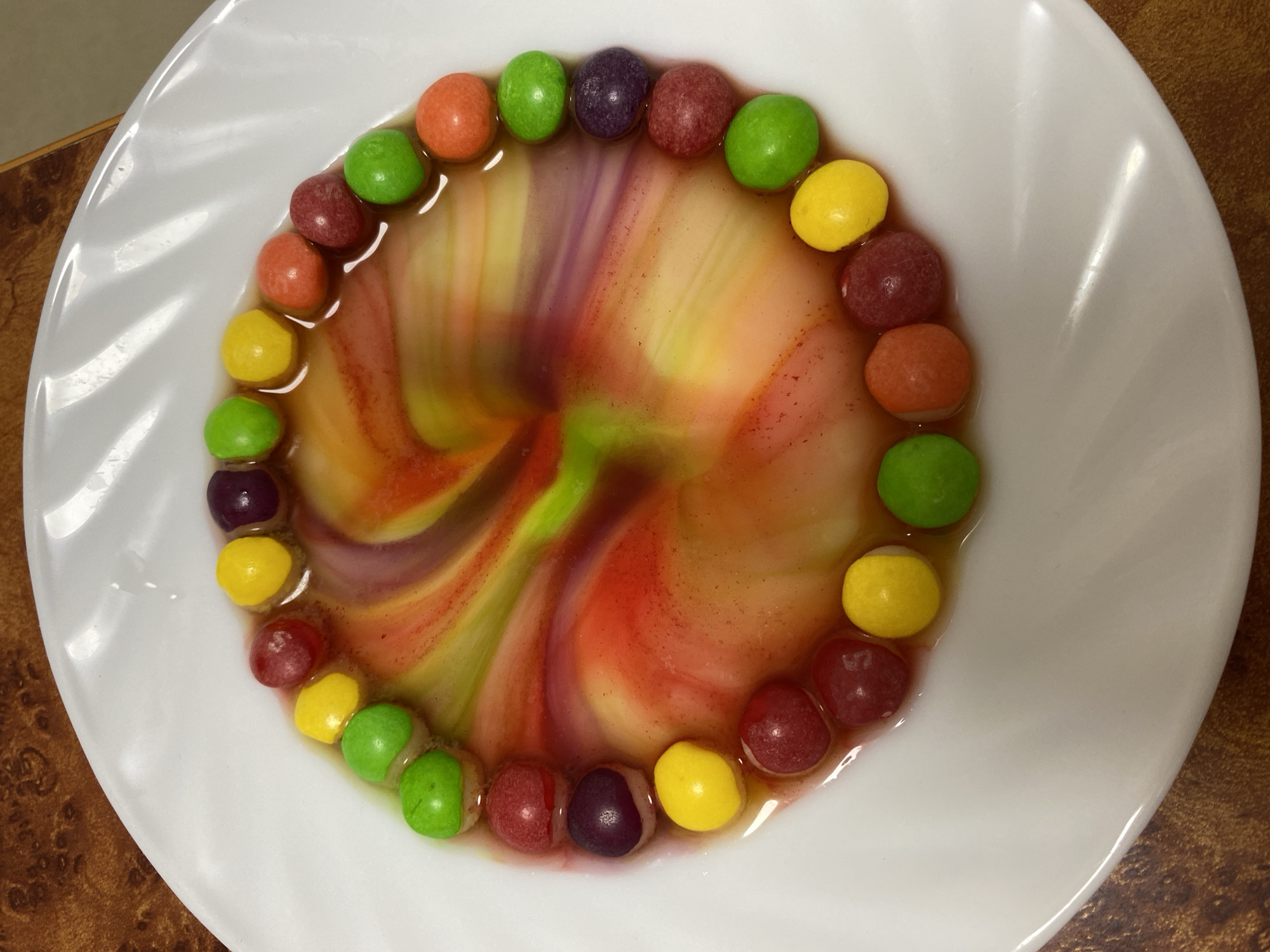 A top-down close-up shows a white plate with a ring of Skittles candies around the edge. The candies are various colors, including green, yellow, orange, red, and purple. Water has been poured onto the plate, dissolving the colored coatings of the Skittles and creating a swirling pattern of dyes in the center of the plate. The background is a wooden surface. No people are visible in the image. No city location is indicated in the image.