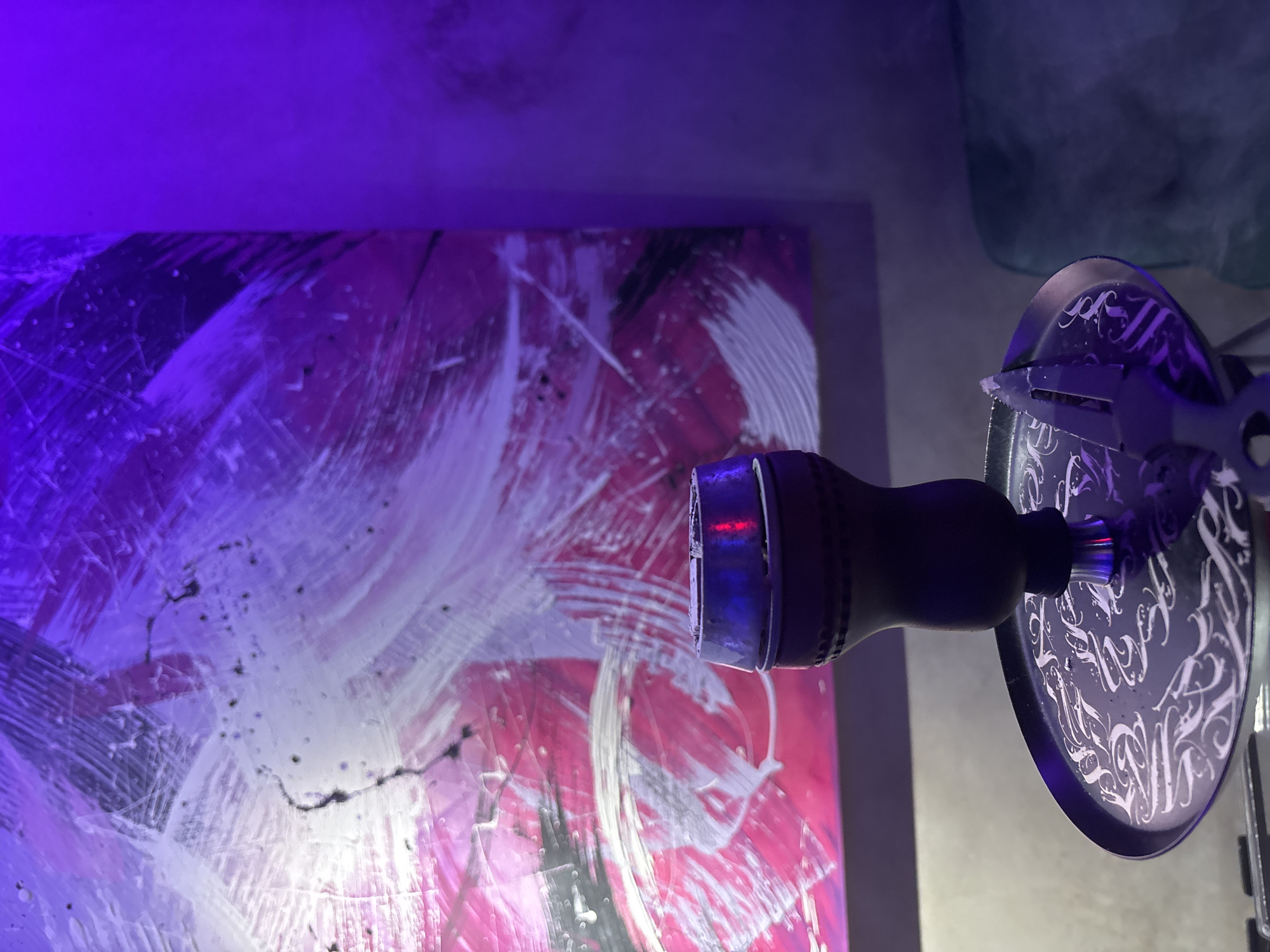 The image depicts a close-up of a hookah pipe with a painting in the background. The painting is abstract, with strokes of white, pink, and black. The hookah is partially visible, focusing on the handle and decorative plate. The decorative plate has a design with curved shapes that resemble stylized script or patterns. The pipe and painting are illuminated by a purple light, with possible reflections or light sources. The image is taken indoors with limited contextual information to pinpoint a specific location. Therefore, determining whether the photo was taken in Kuta Selatan, Indonesia, is impossible without additional context.