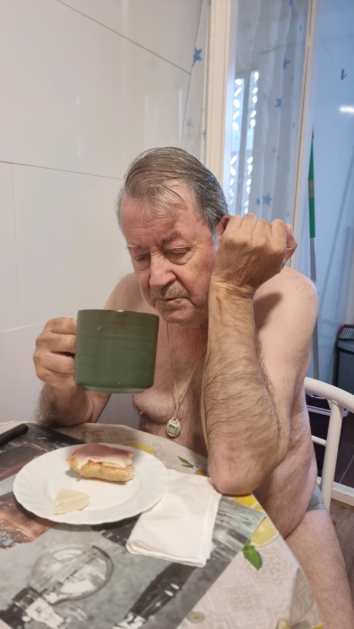 An older man is seated at a table, holding a green mug in his left hand and resting his right elbow on the table, his head slightly tilted downwards and eyes closed. He is shirtless, wearing what appears to be grey underwear, and has a necklace with a white pendant. In front of him on the table is a white plate with a small sandwich topped with ham and cheese, and a triangular piece of cheese. A white napkin is folded to the right of the plate. The table has a patterned tablecloth featuring lemons. The background shows a white tiled wall and a window with a sheer curtain. The scene appears to be indoors, likely in a residential setting in Sant Joan Les Fonts, Spain.